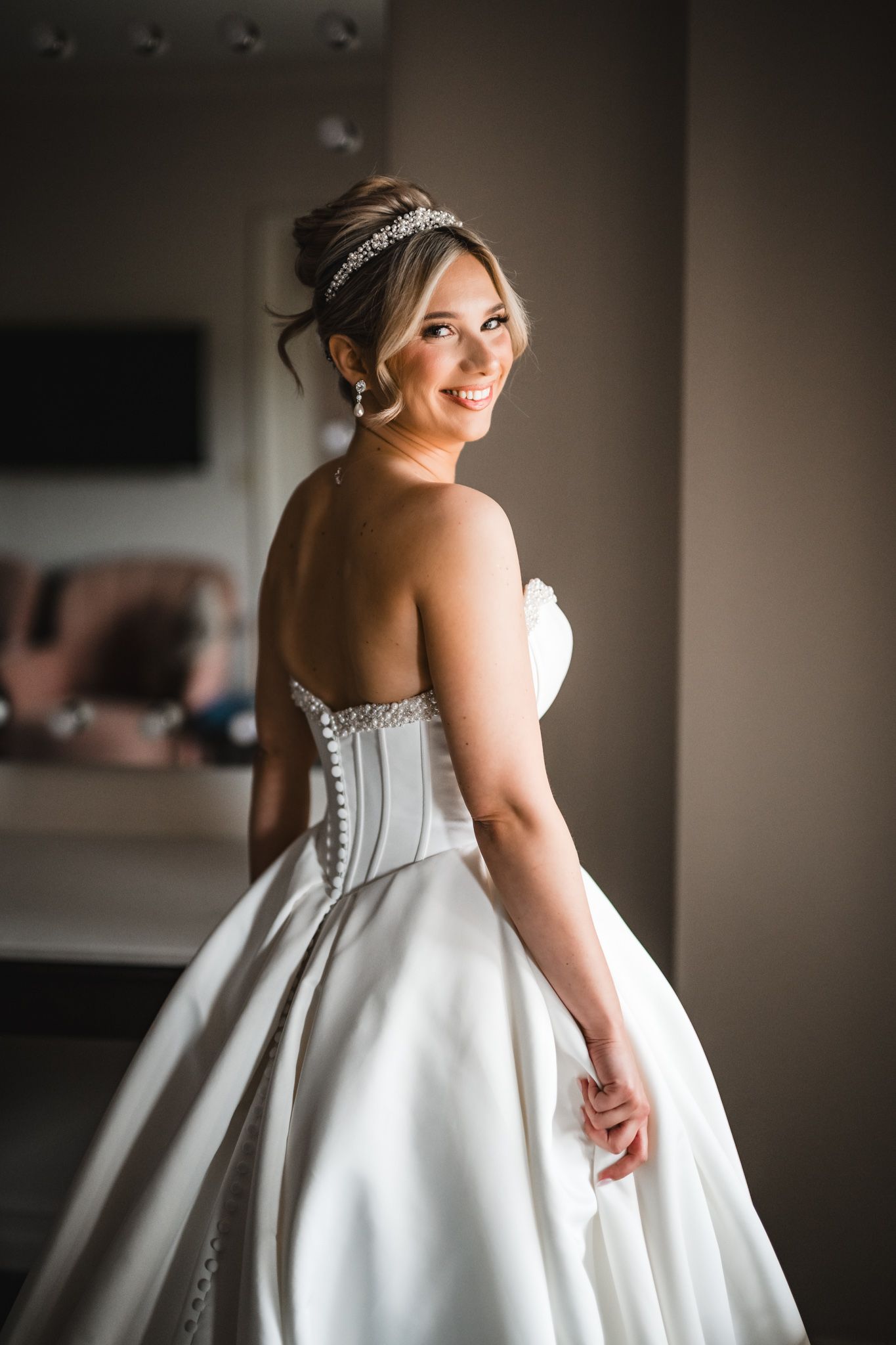 Smiling bride in strapless beaded ballgown with crystal tiara posing over shoulder in bridal suite.