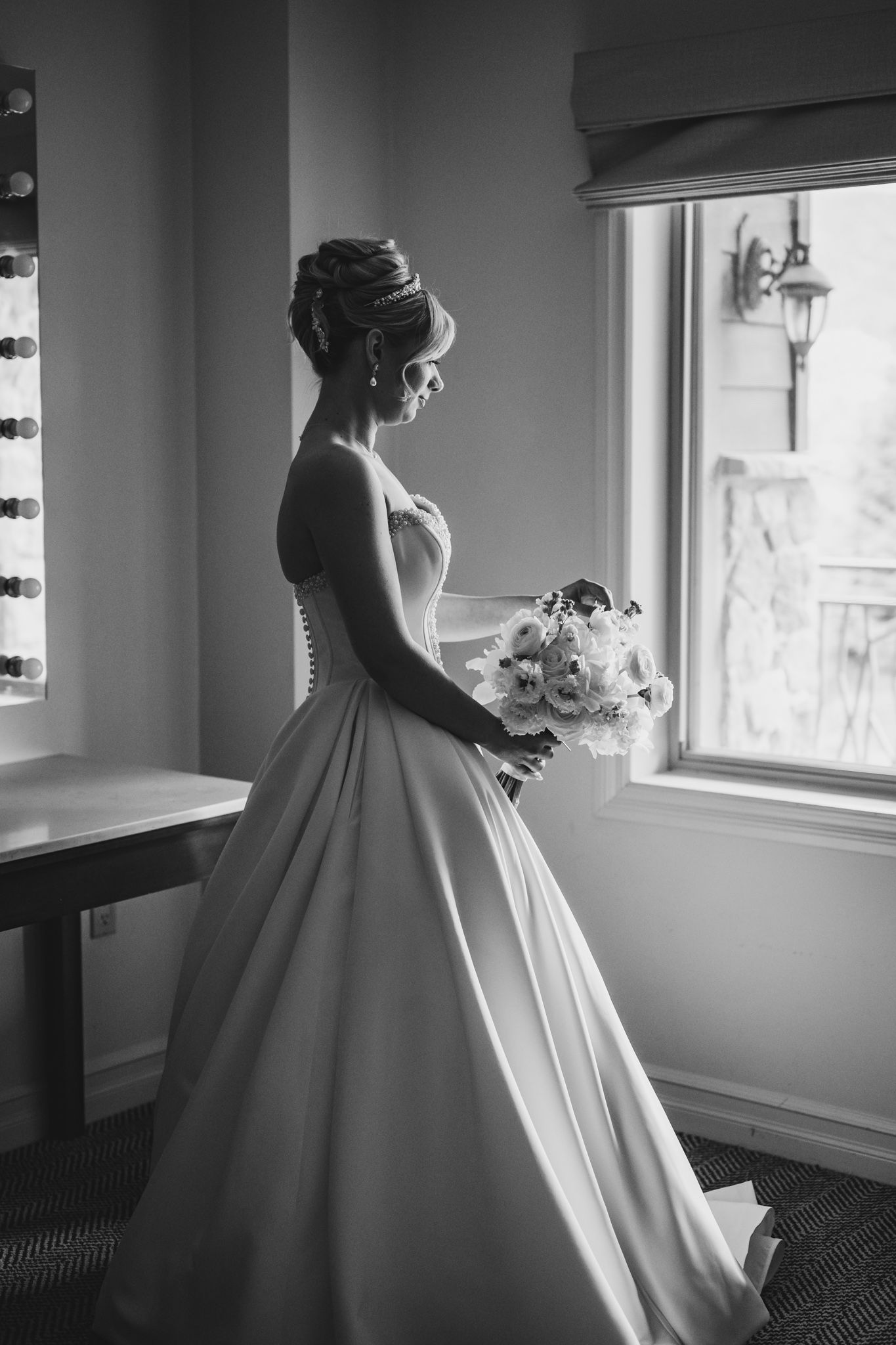 Black and white photo of bride in strapless ball gown holding bouquet by window.