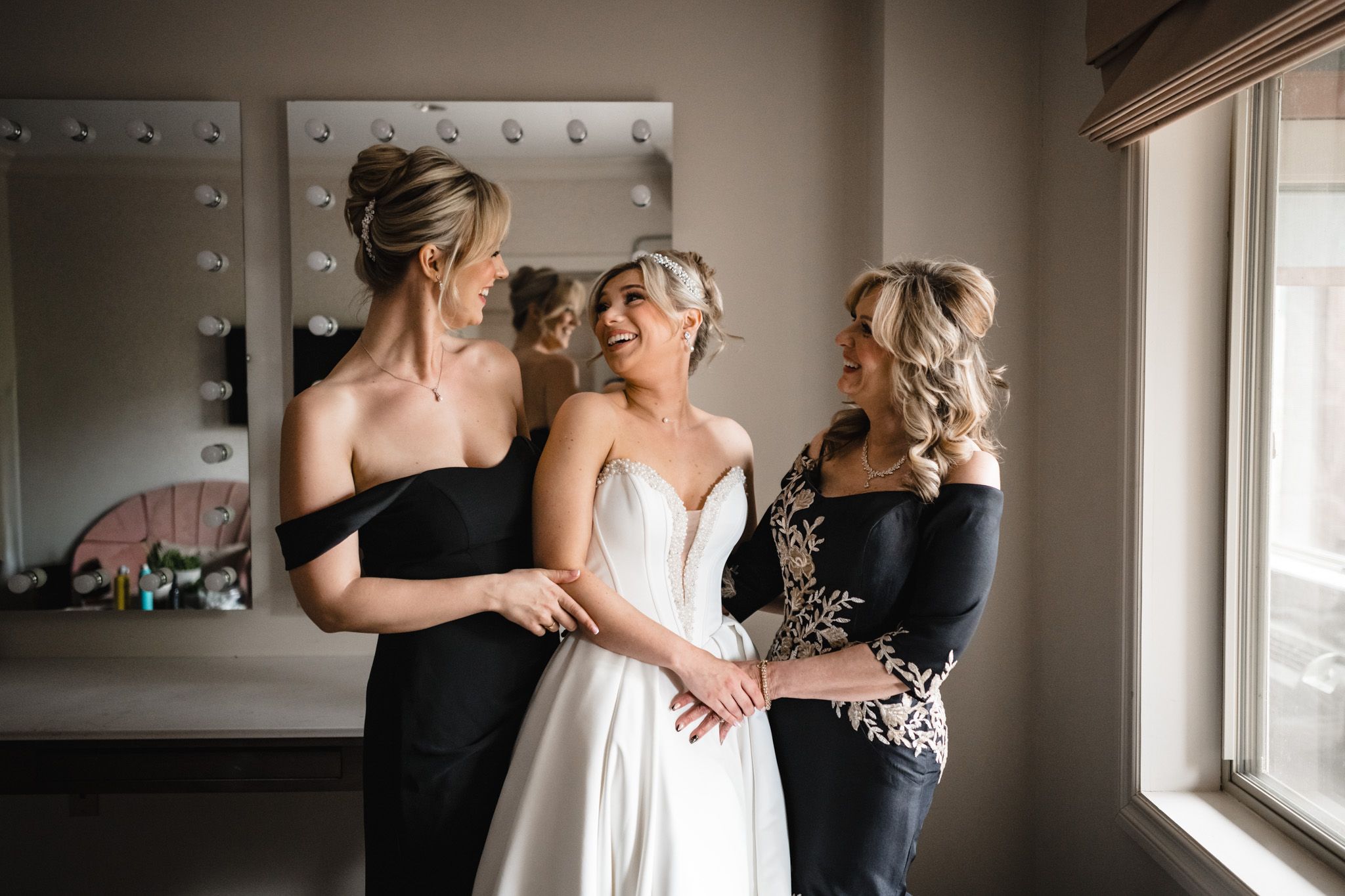 Bride in white gown smiles with two women in black dresses near a mirror during wedding preparations.