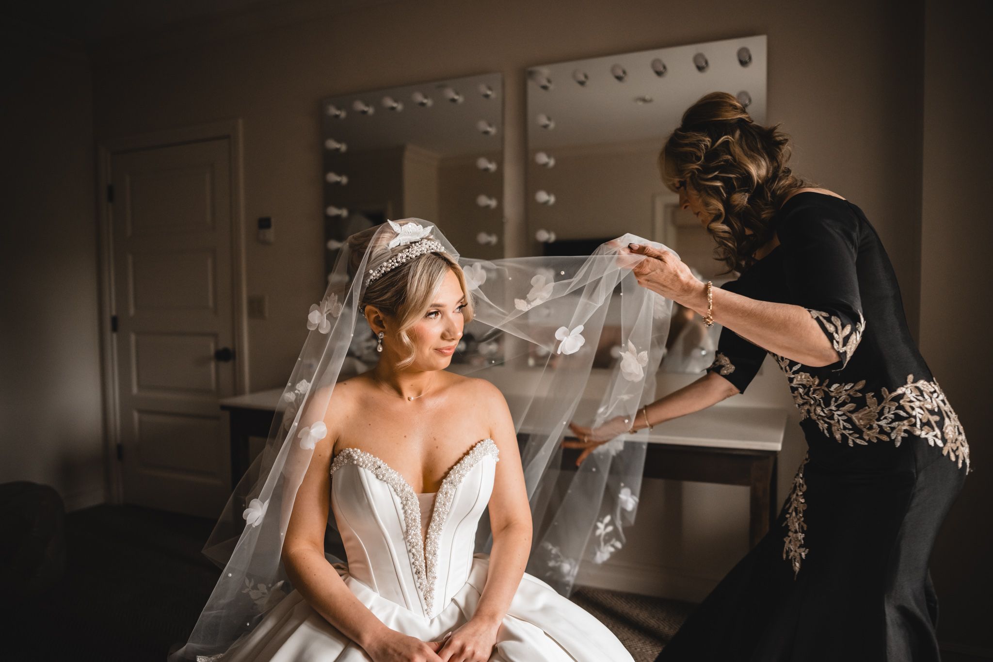 Bridesmaid arranging floral veil on seated bride in strapless beaded wedding gown in dressing room.