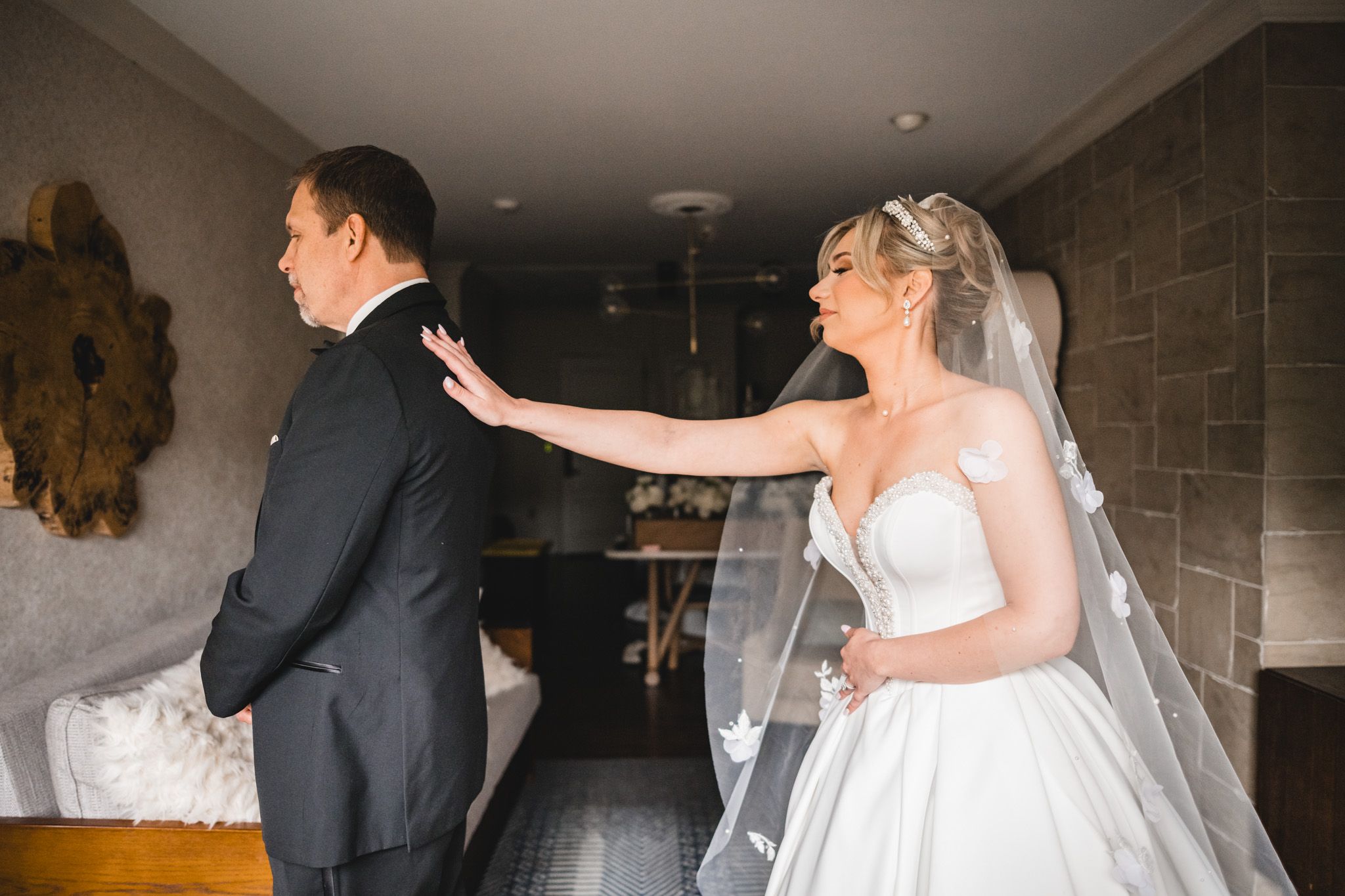 Bride in veil and ball gown tapping groom's shoulder during emotional first look reveal in hotel room.