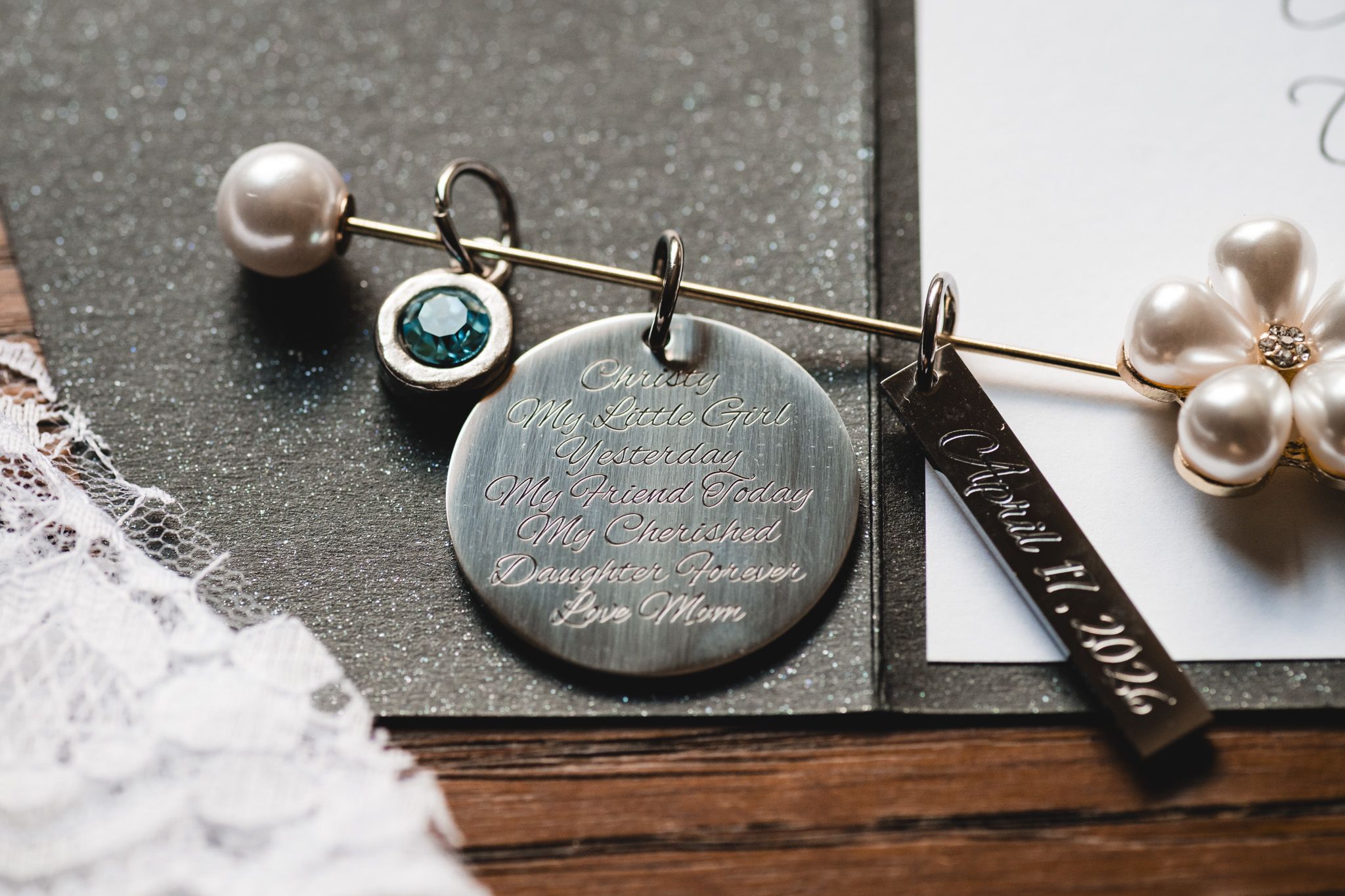 Engraved silver wedding charm pendant with pearl and emerald brooch pins on dark surface, sentimental bridal jewelry accessories.