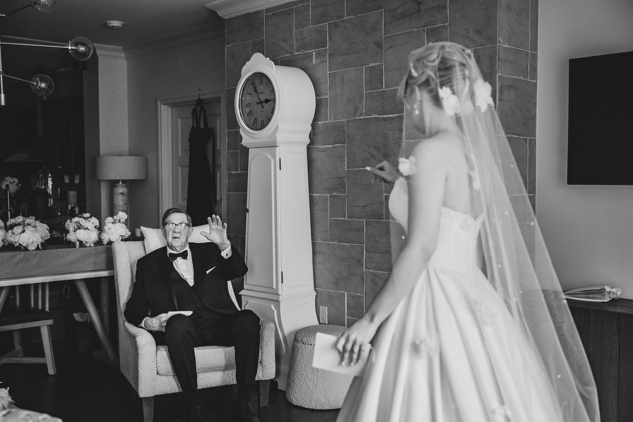 Black and white photo of a groom seated in a chair reacting as his bride in a veil approaches him.
