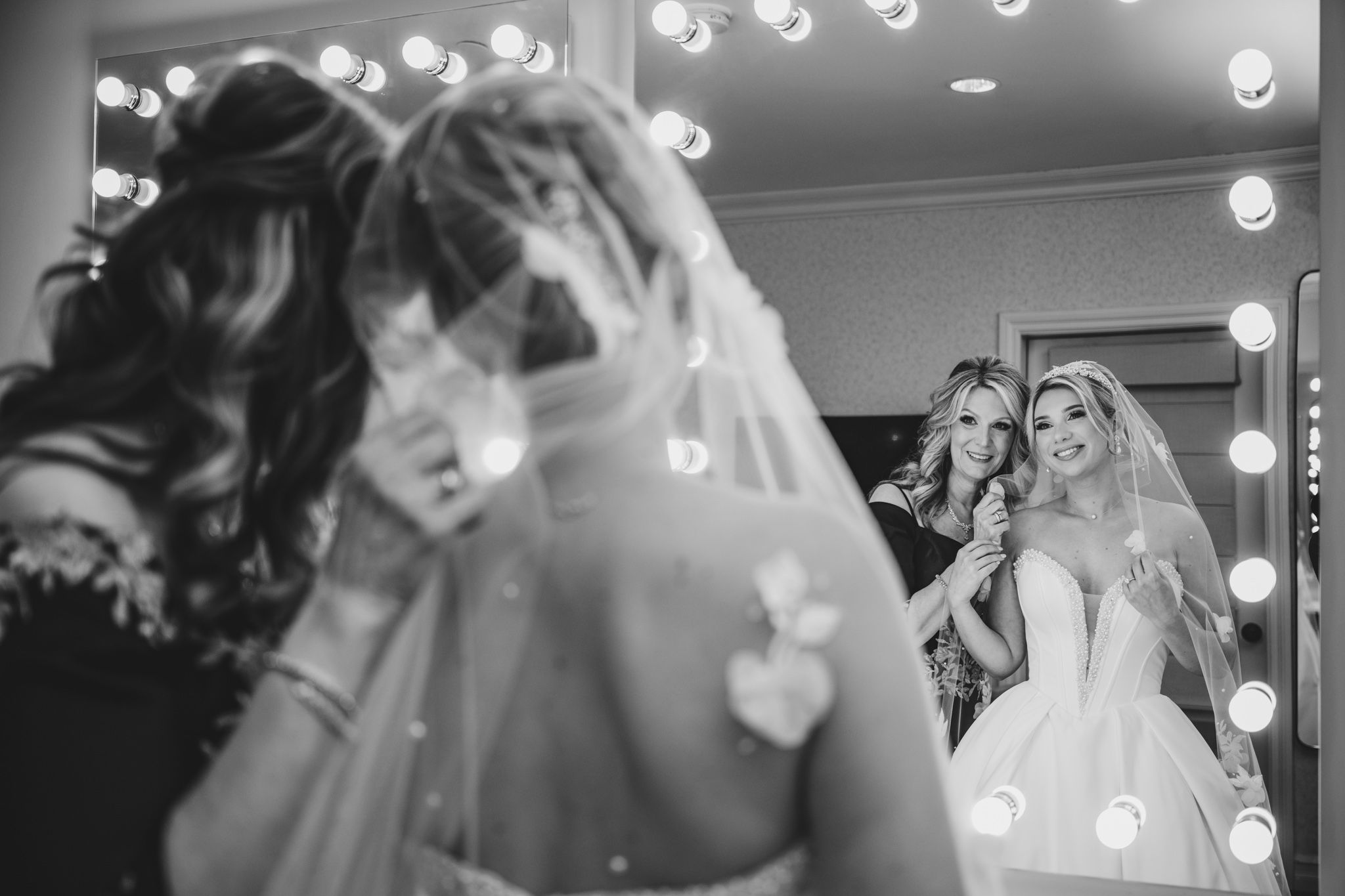 Black and white photo of a bride in a veil and gown smiling with a bridesmaid reflected in a vanity mirror.