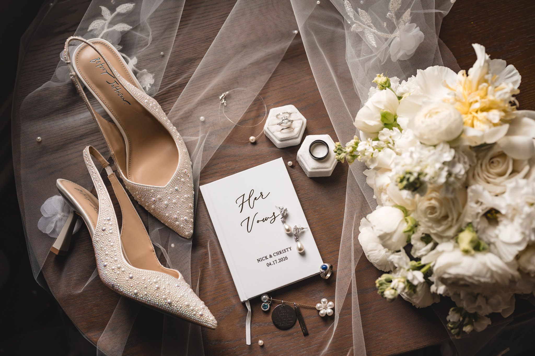 Bridal flat lay featuring pearl heels, vow book, ring box, perfume, and white floral bouquet on wooden surface.