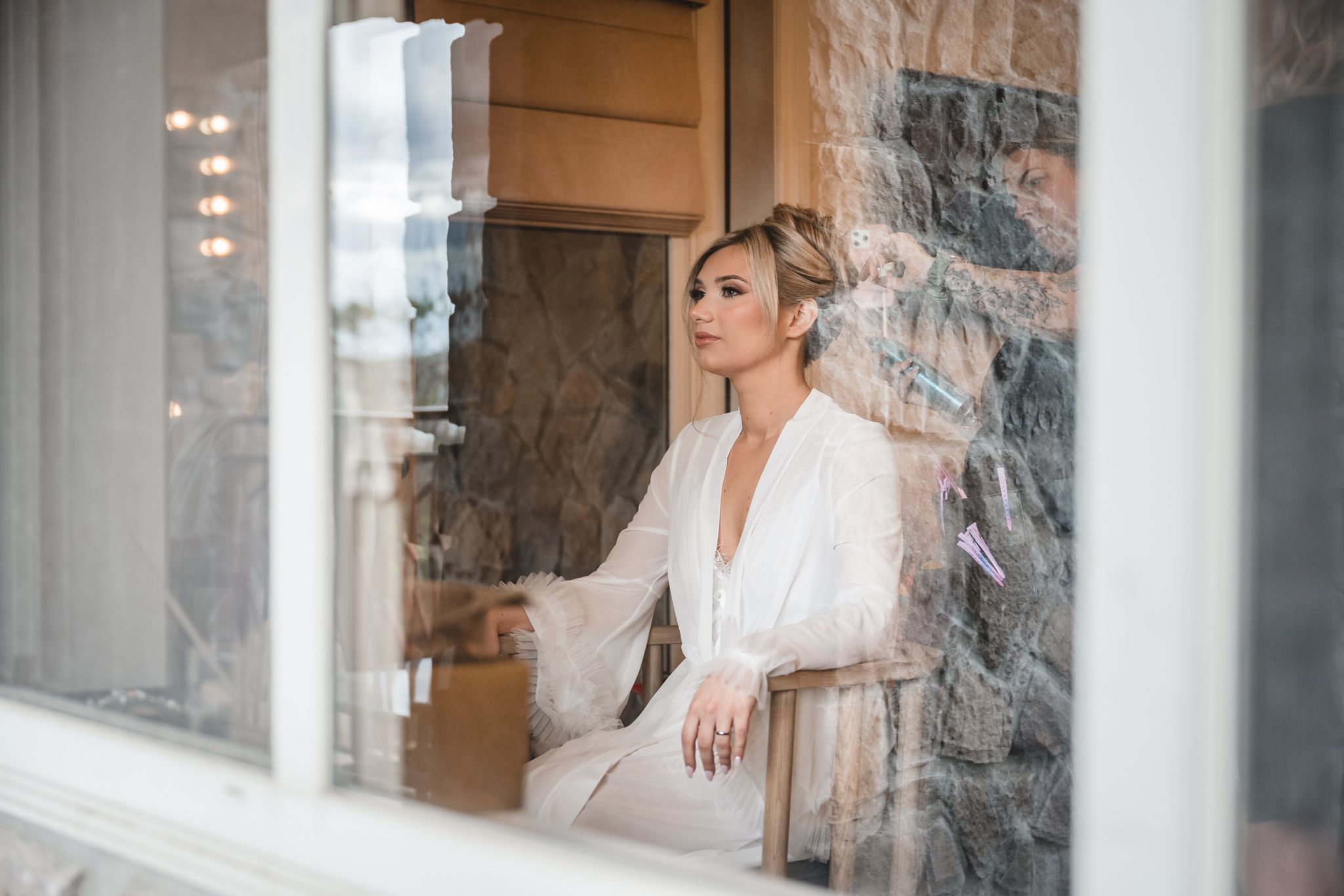 Elegant bride in white lace robe sitting thoughtfully by a window with stone wall backdrop.
