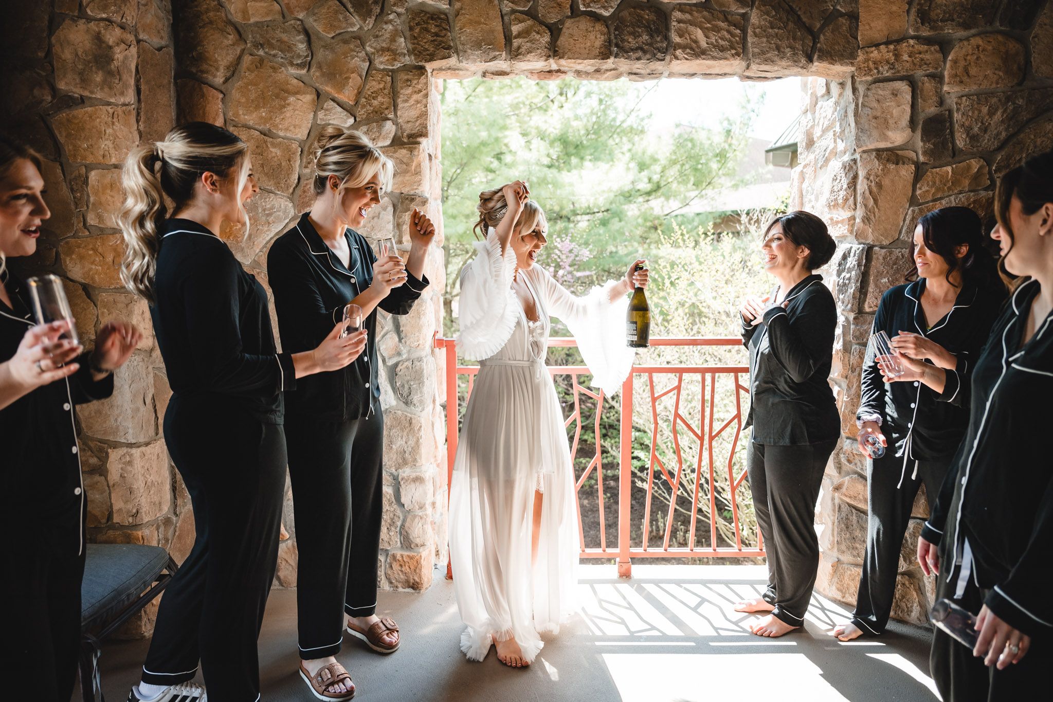 Bride in white gown walks through stone arch as bridesmaids in black pajamas cheer.