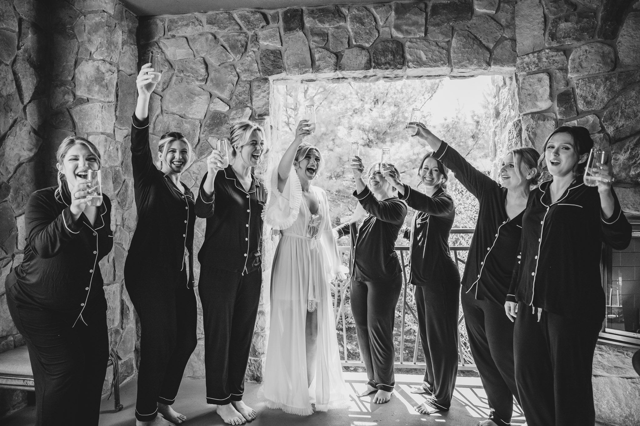 Black and white photo of bride and bridesmaids in pajamas raising arms joyfully under stone arch.