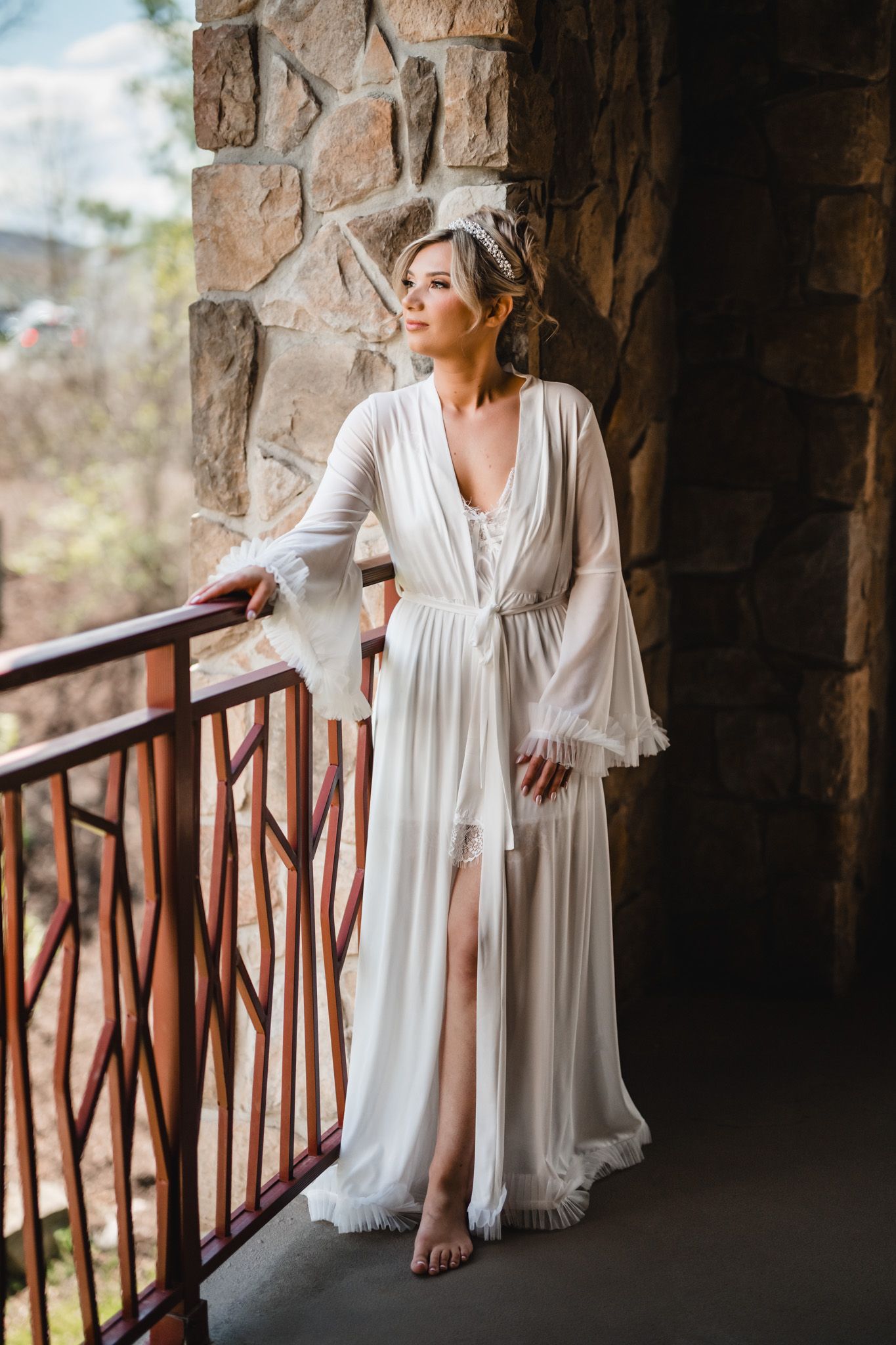 Bride in flowing white chiffon robe with bell sleeves and crystal tiara standing on stone balcony.