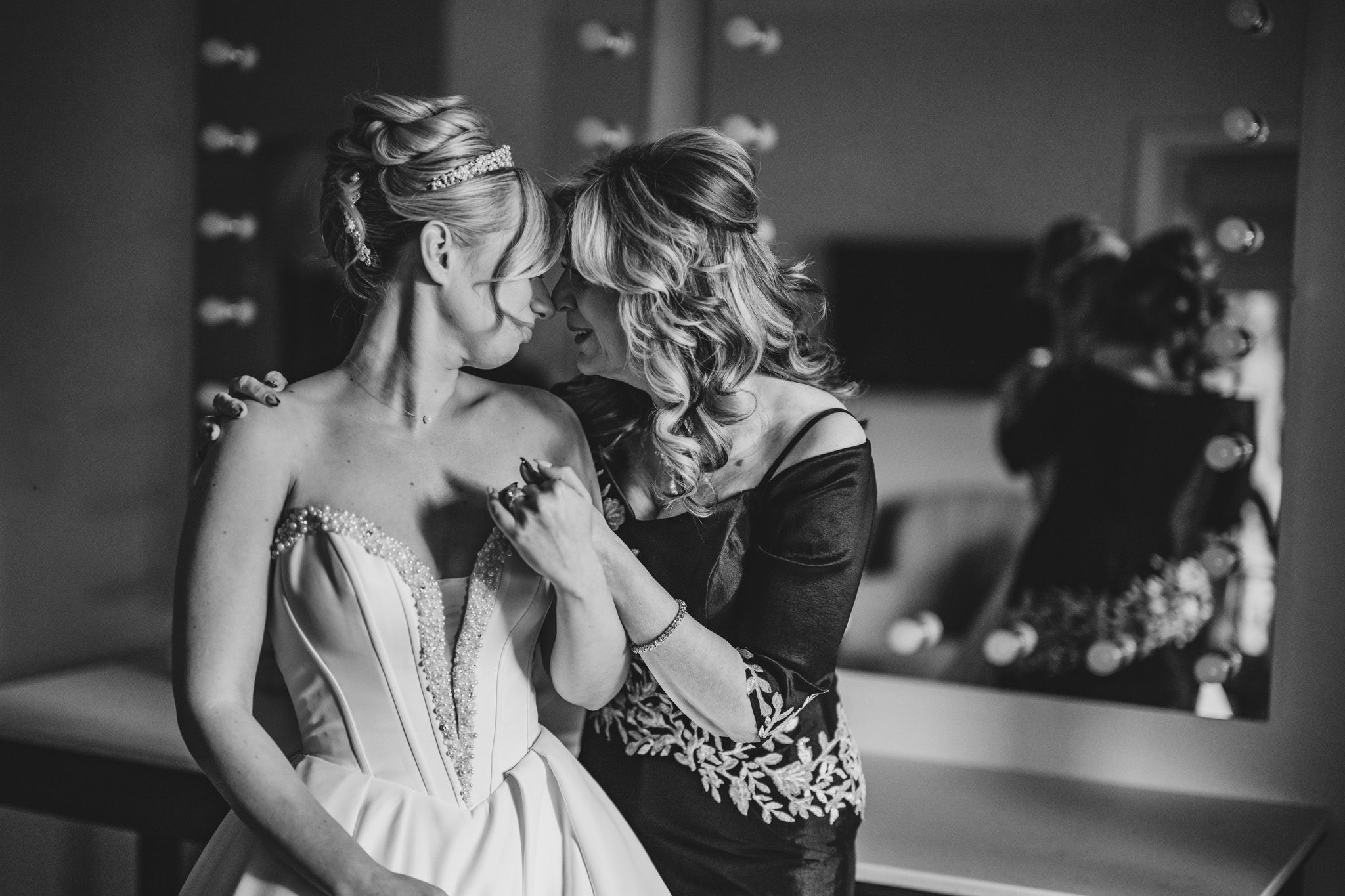 Black and white photo of a bride getting her dress fastened by a woman in front of a lit vanity mirror.