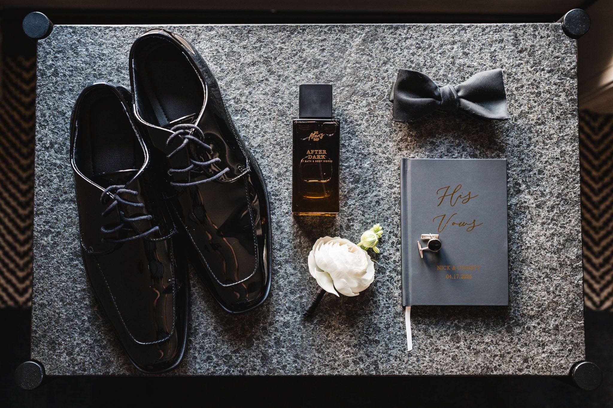 Groom wedding day flat lay with black patent shoes, bow tie, cologne, boutonniere and vow book.