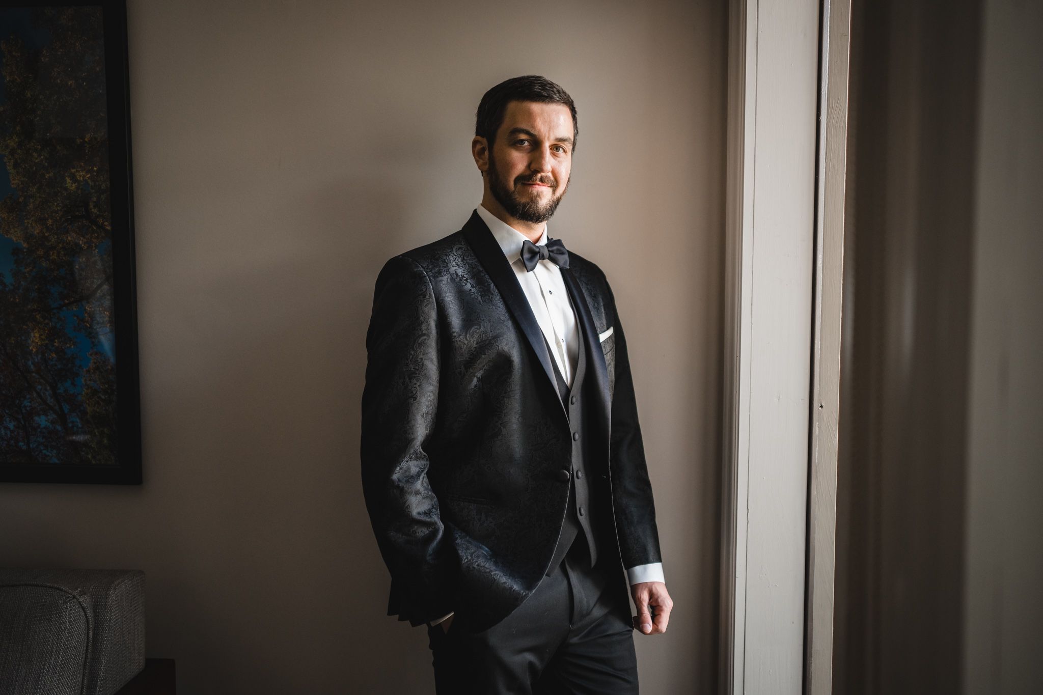 Groom in black tuxedo with bow tie posing confidently by a window in soft natural light.