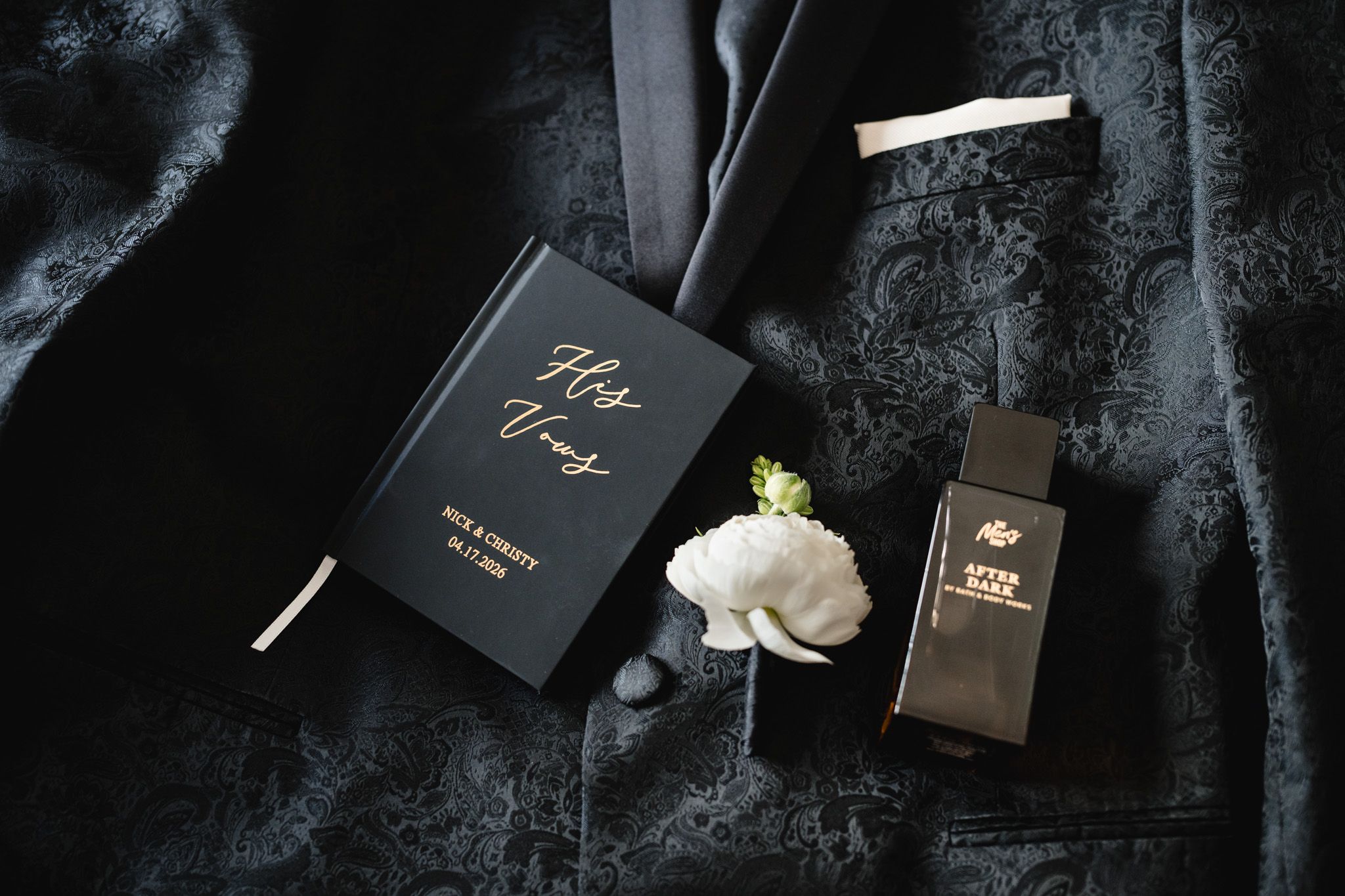Groom's wedding flat lay with black vow book, white boutonniere, cologne, and dark suit jacket.