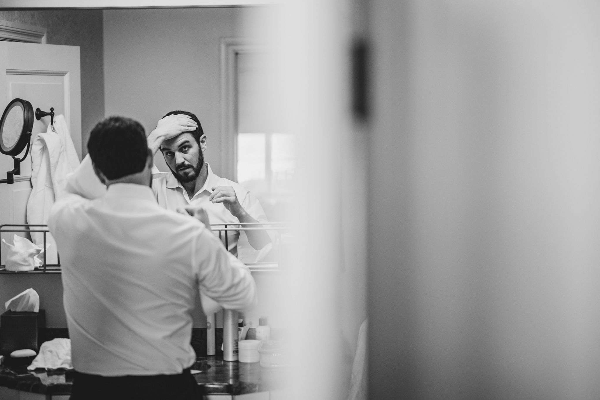 Groom in white dress shirt adjusts his tie while looking in the mirror before his wedding.