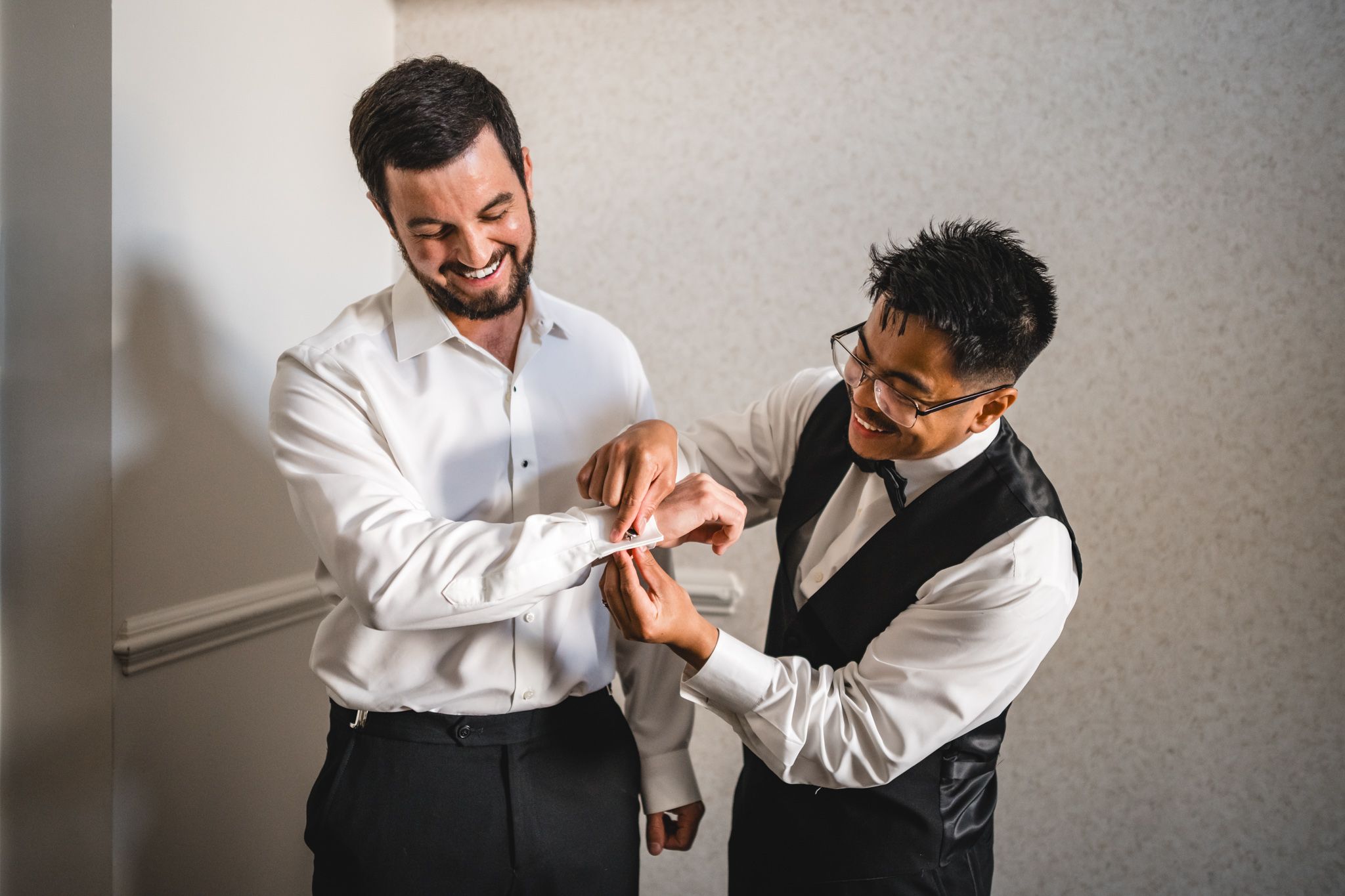 Smiling groom getting cufflinks fastened by groomsman in white dress shirt before wedding ceremony.