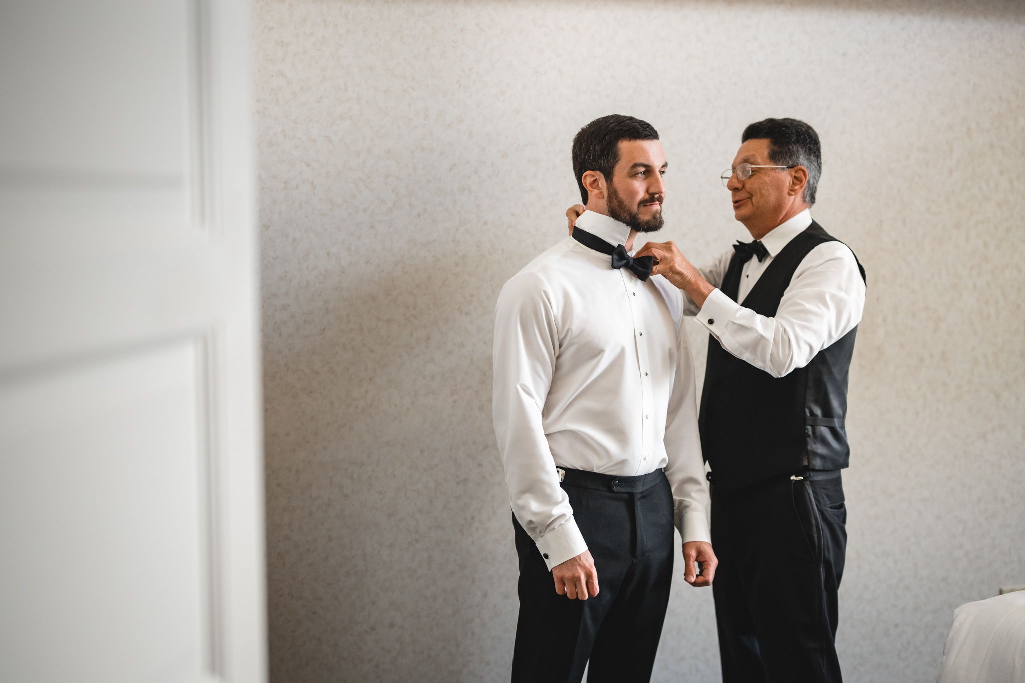 Father helping groom adjust bow tie during wedding day preparations in a bright room.