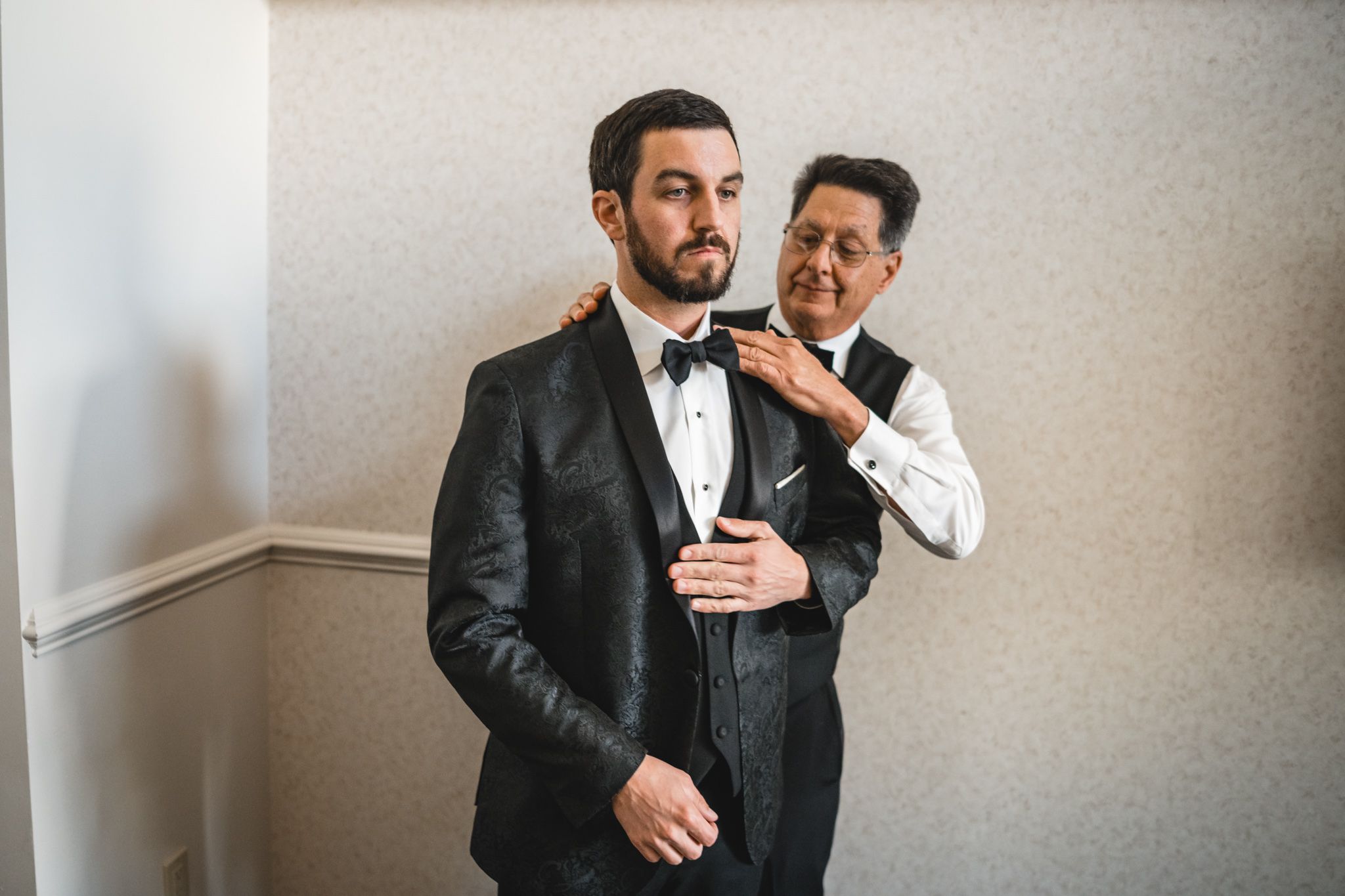 Groom in black tuxedo having bow tie adjusted by father or groomsman before wedding ceremony.