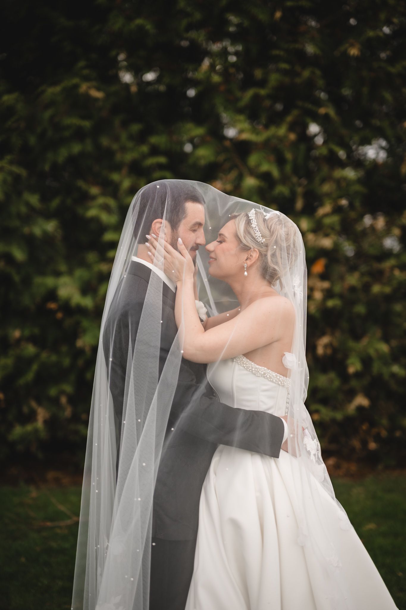 Bride and groom share a romantic moment under a pearl-dotted veil outdoors on their wedding day.