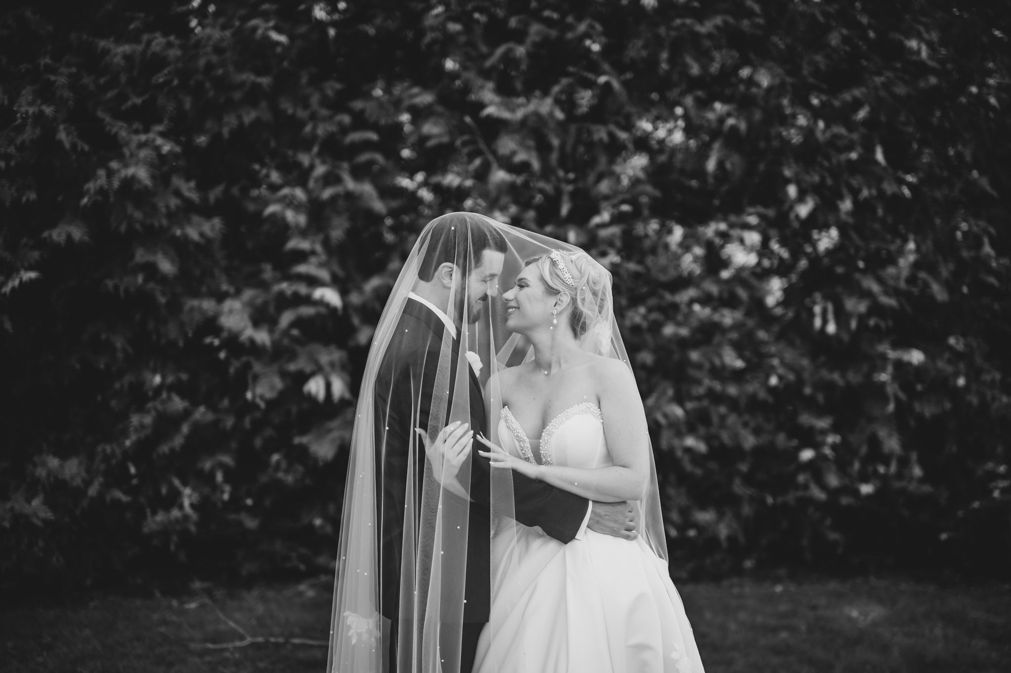 Bride and groom sharing a romantic kiss under a flowing white veil in a black and white wedding portrait.