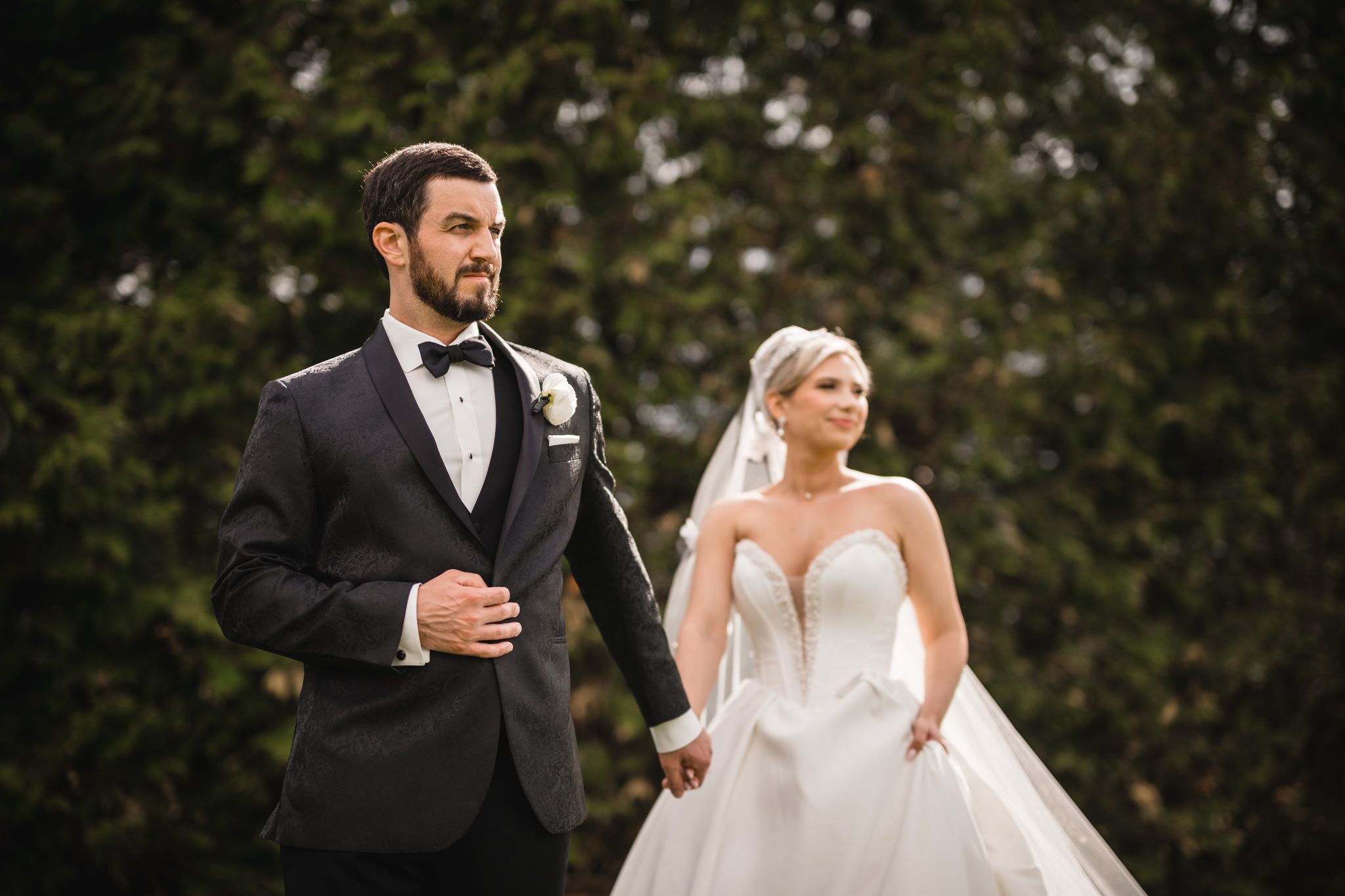 Elegant bride and groom holding hands outdoors, with groom in tuxedo and bride in a ball gown with veil.