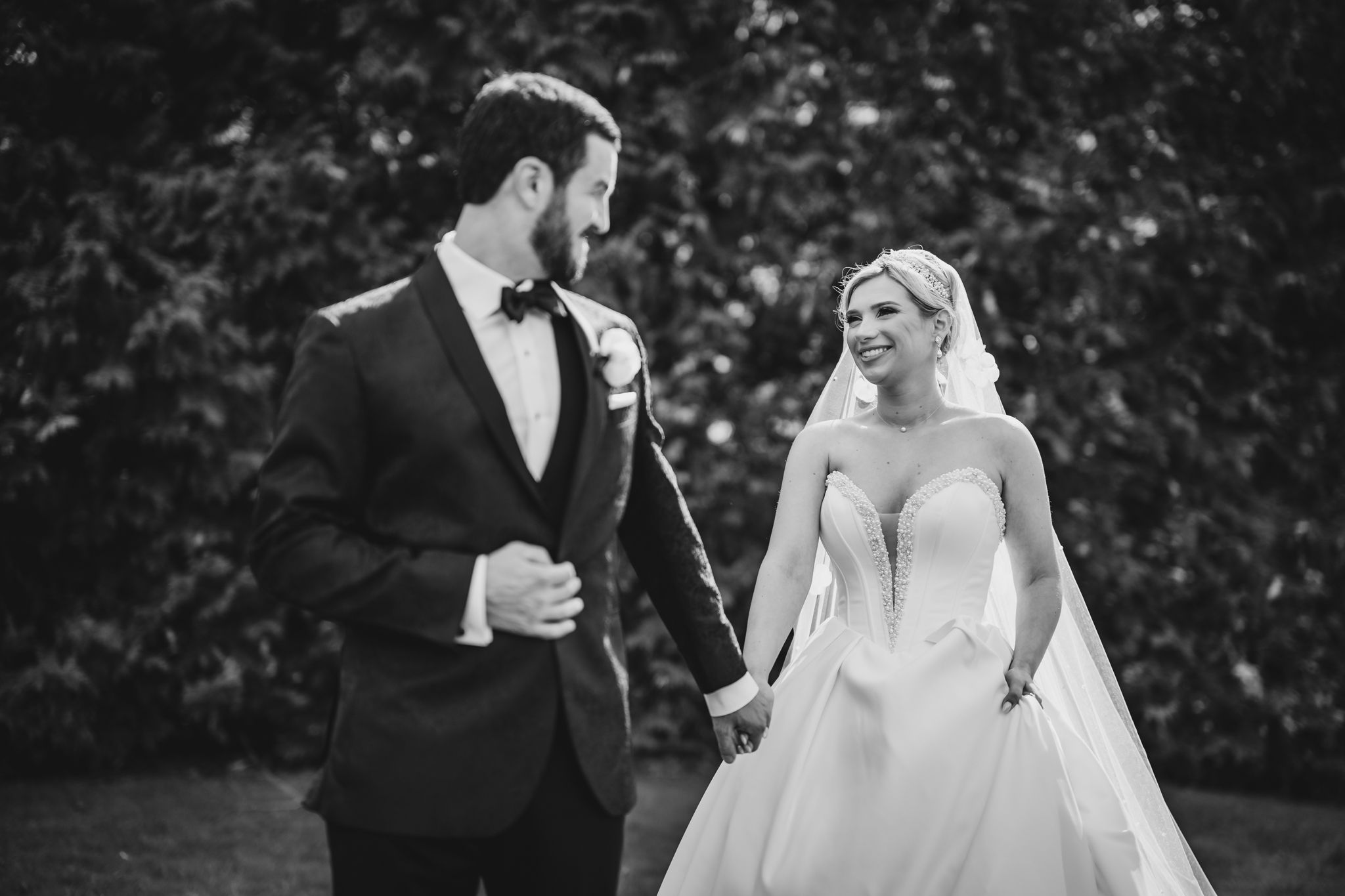 Black and white photo of a newlywed couple holding hands and smiling at each other in a garden.