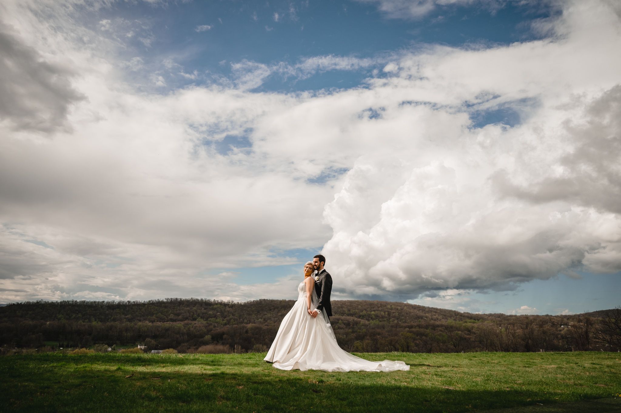 Bride and groom stand together on lush green lawn with stunning mountain landscape and dramatic cloudy sky backdrop.