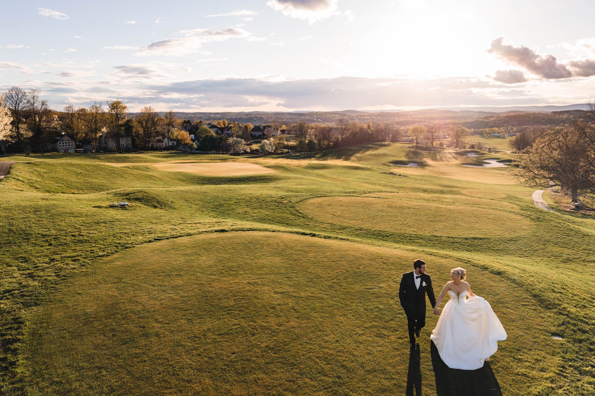 Aerial drone view of bride and groom walking hand in hand across a lush golf course at golden hour.