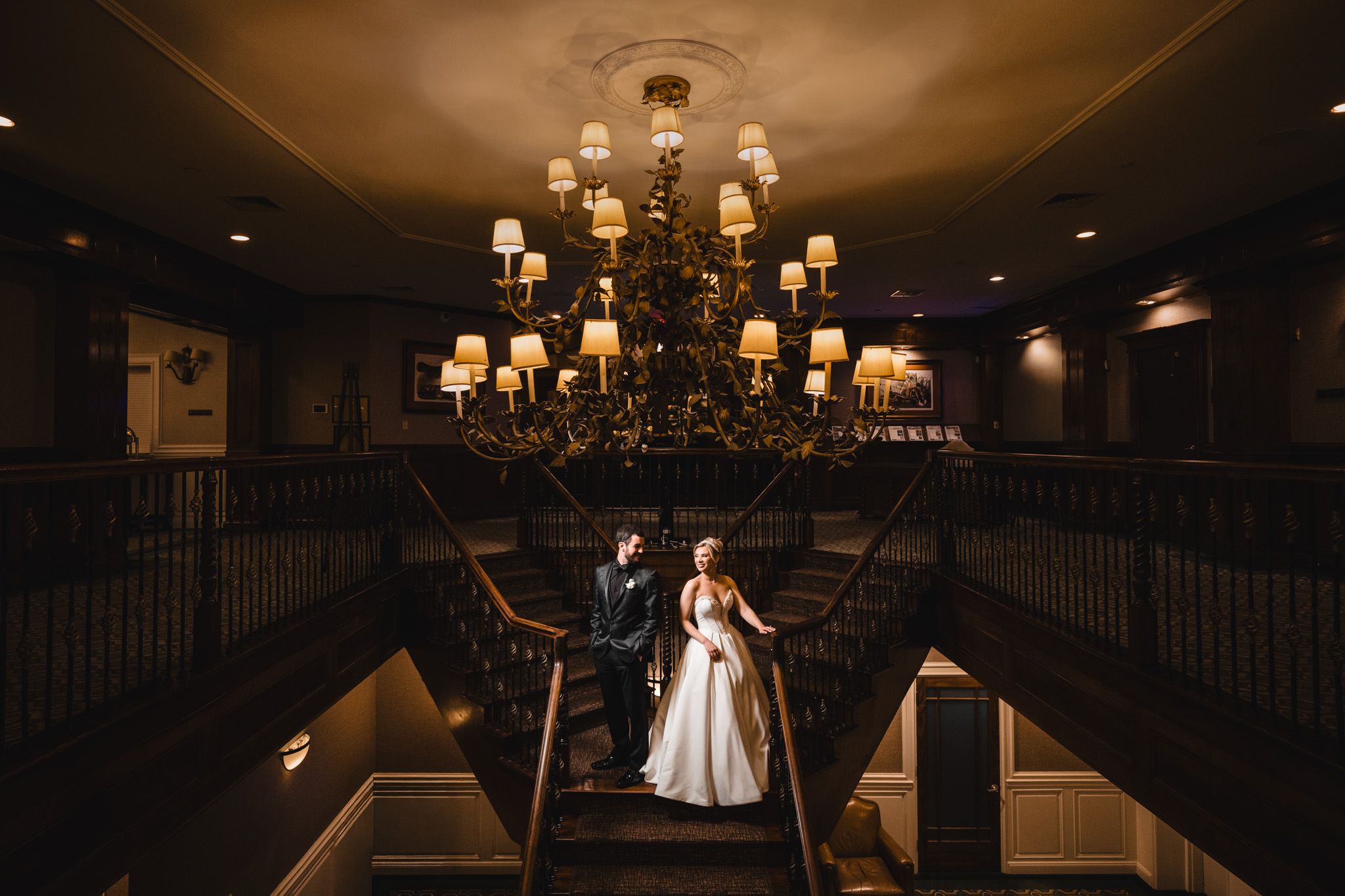 Bride and groom descend a grand staircase beneath an elegant chandelier in a dimly lit venue.