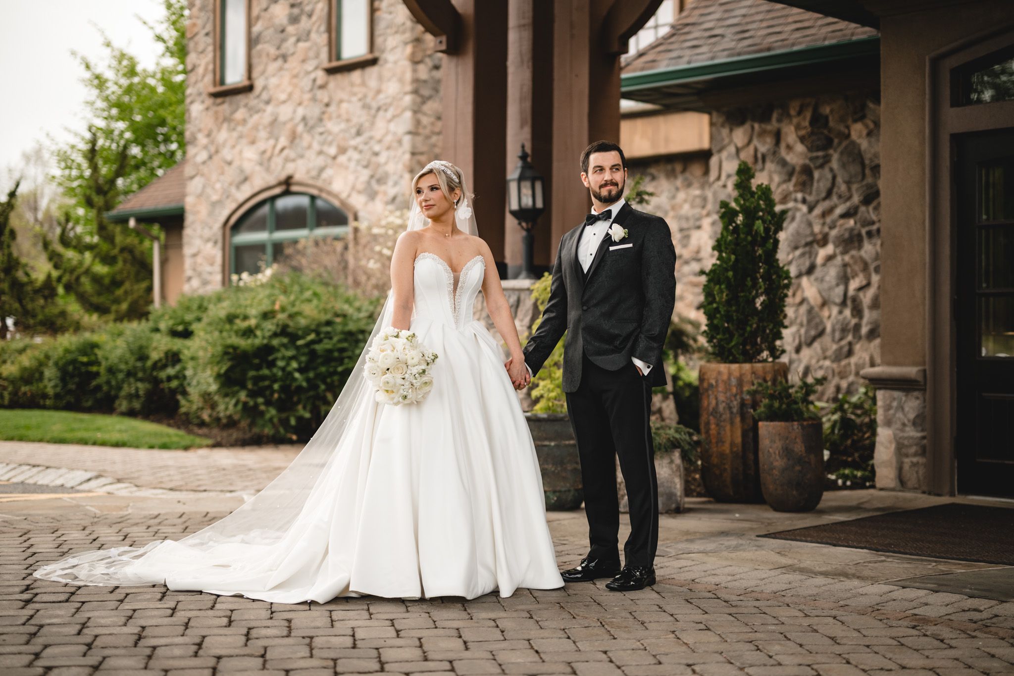 Bride and groom portrait outside stone venue, bride in ballgown with bouquet, groom in black tux.