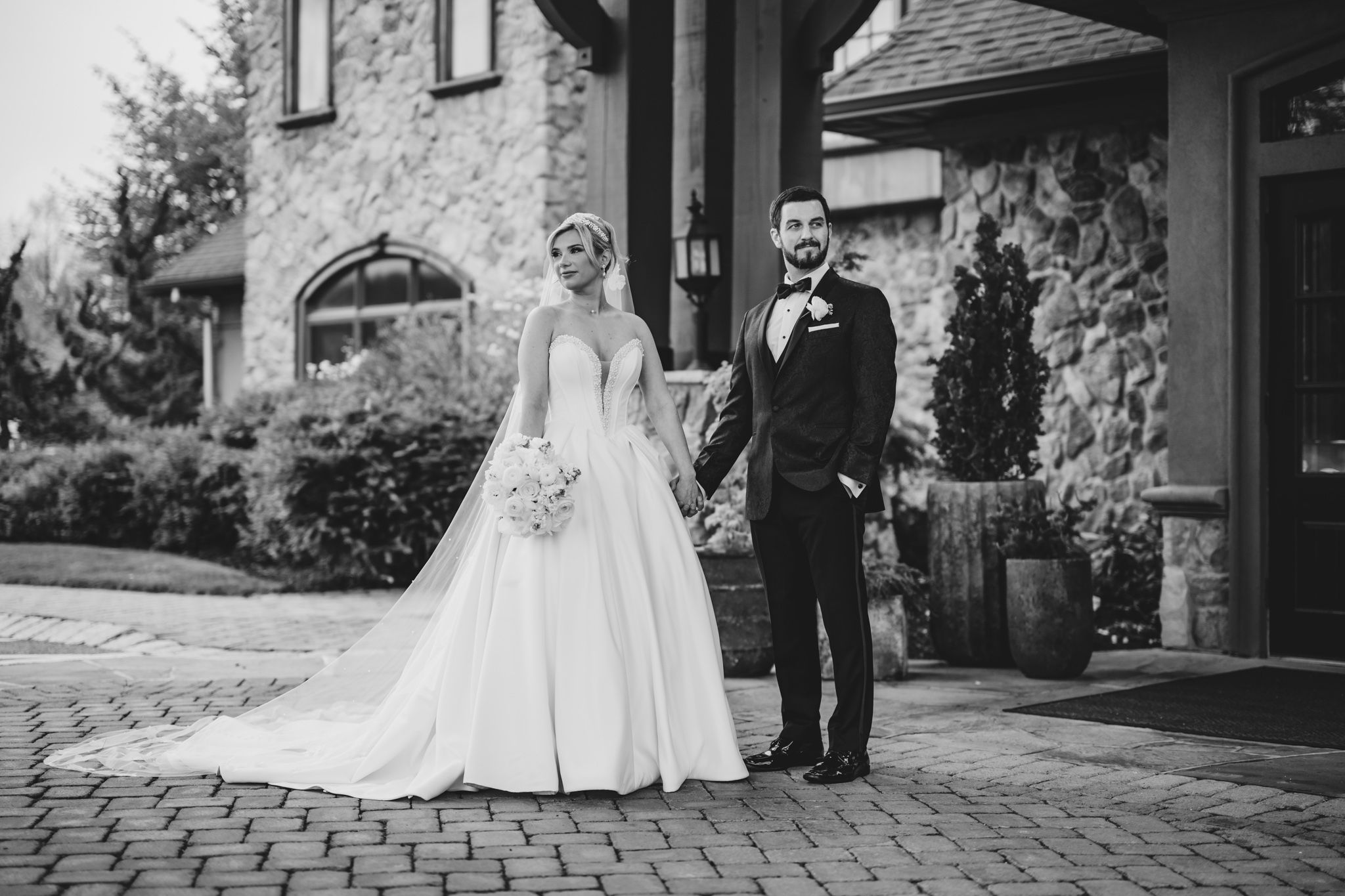 Black and white portrait of bride and groom holding hands outside a rustic stone wedding venue.