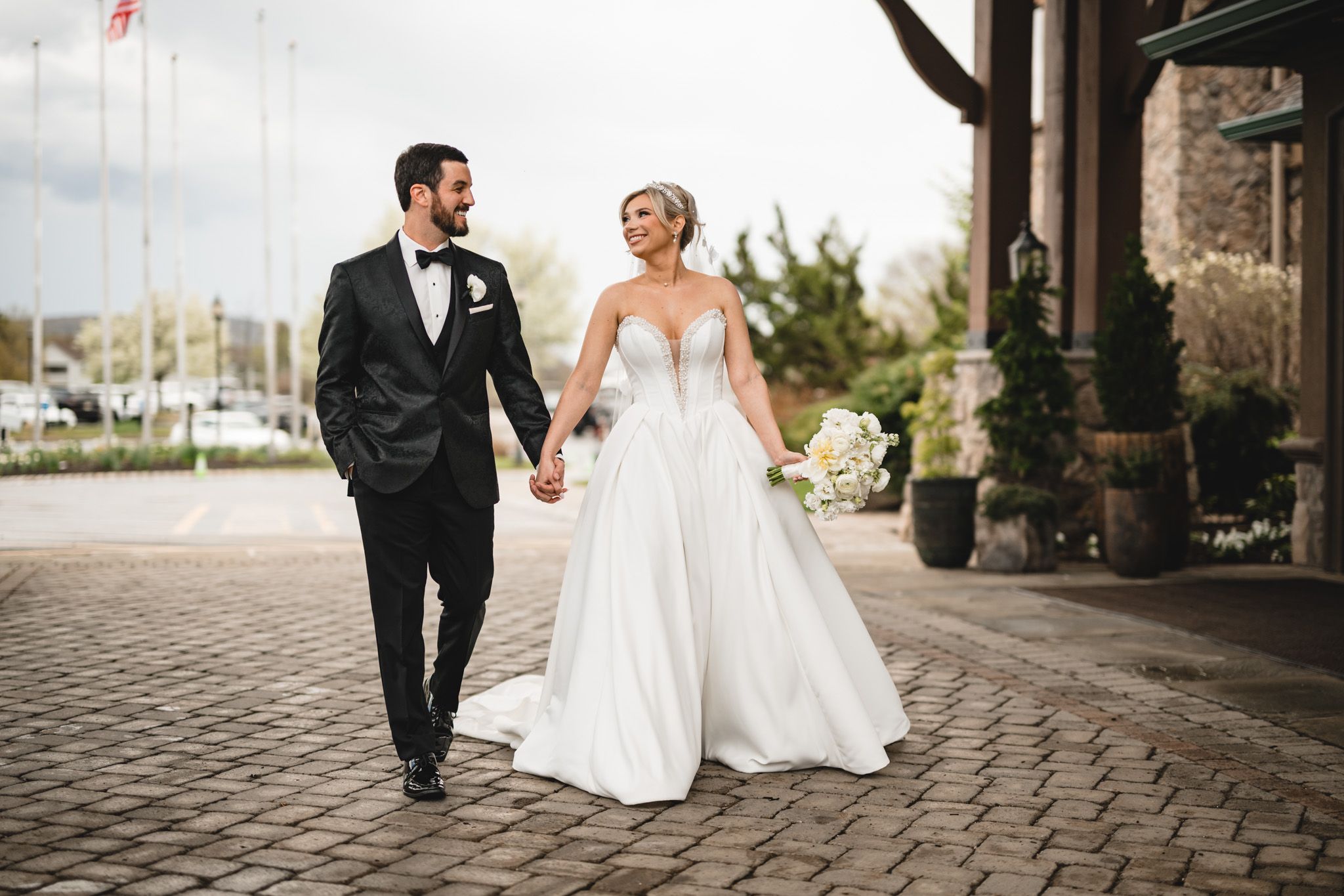 Bride and groom walk hand-in-hand on cobblestone path, she in a white gown, he in a black tuxedo.