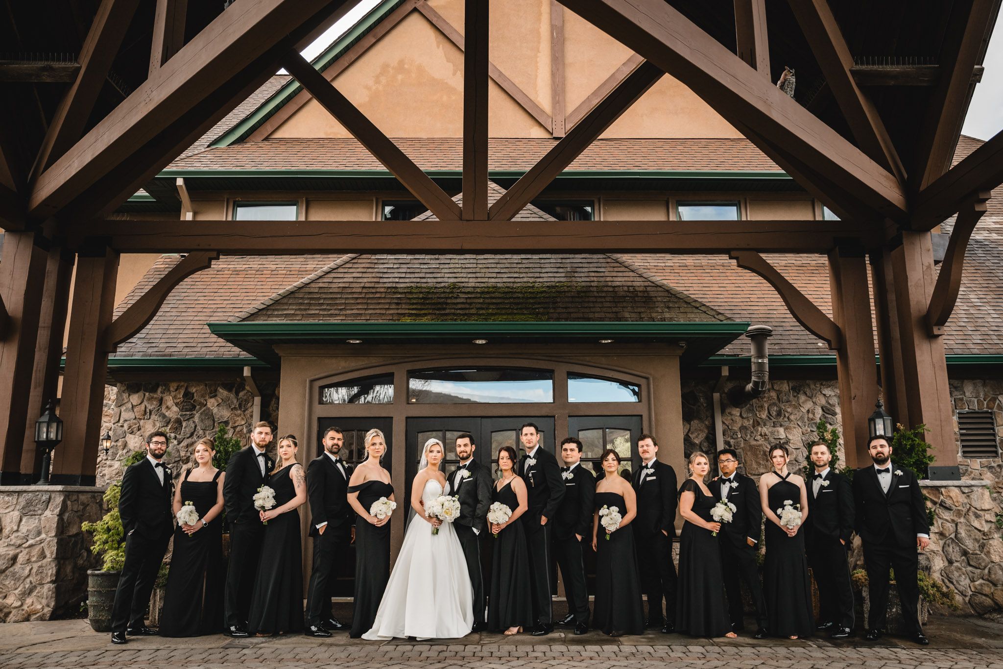Wedding party in all-black attire poses with bride and groom under a wooden venue entrance.