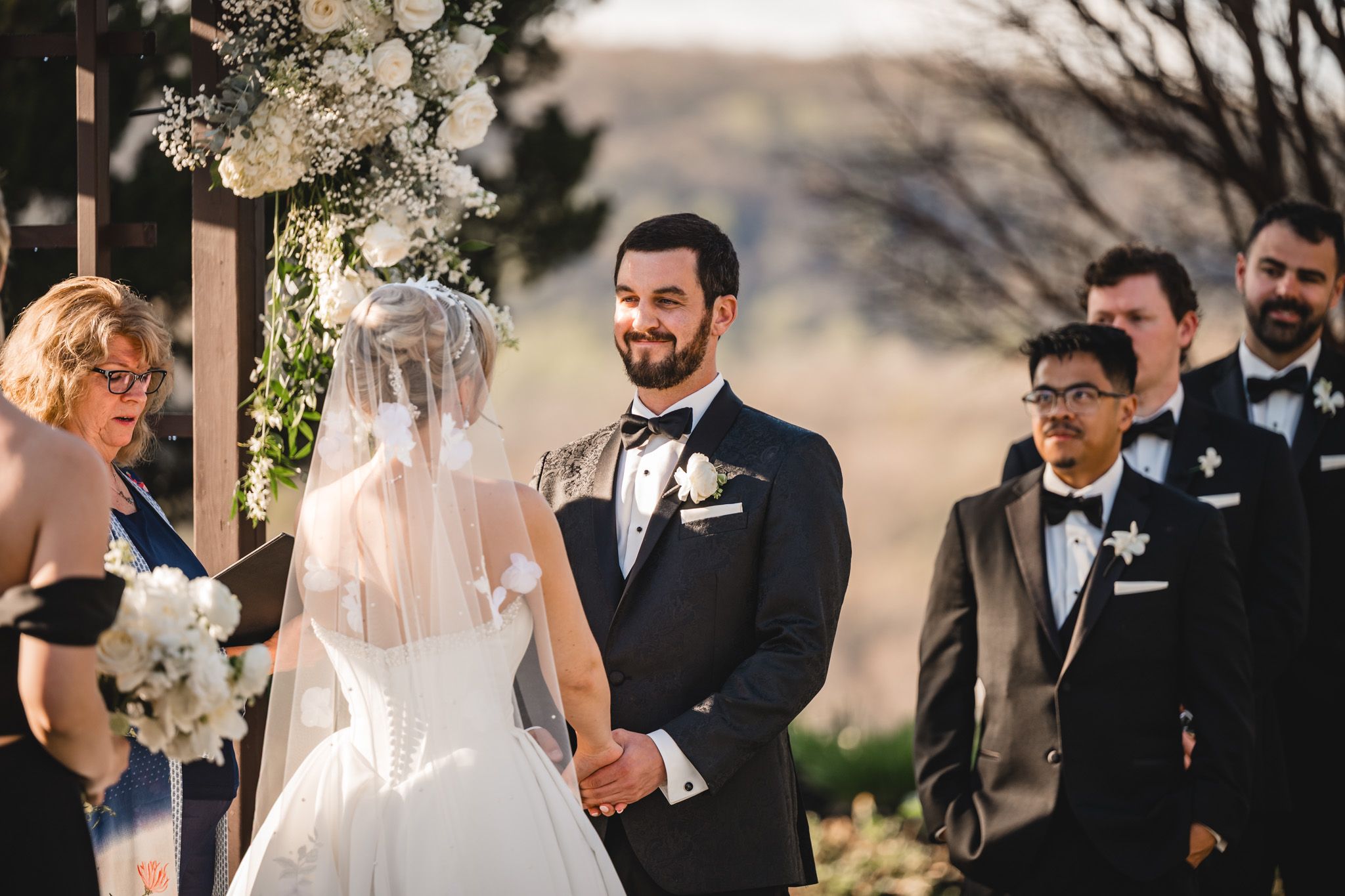 Groom smiles at bride during outdoor wedding ceremony with groomsmen and officiant beside rustic wooden arch.