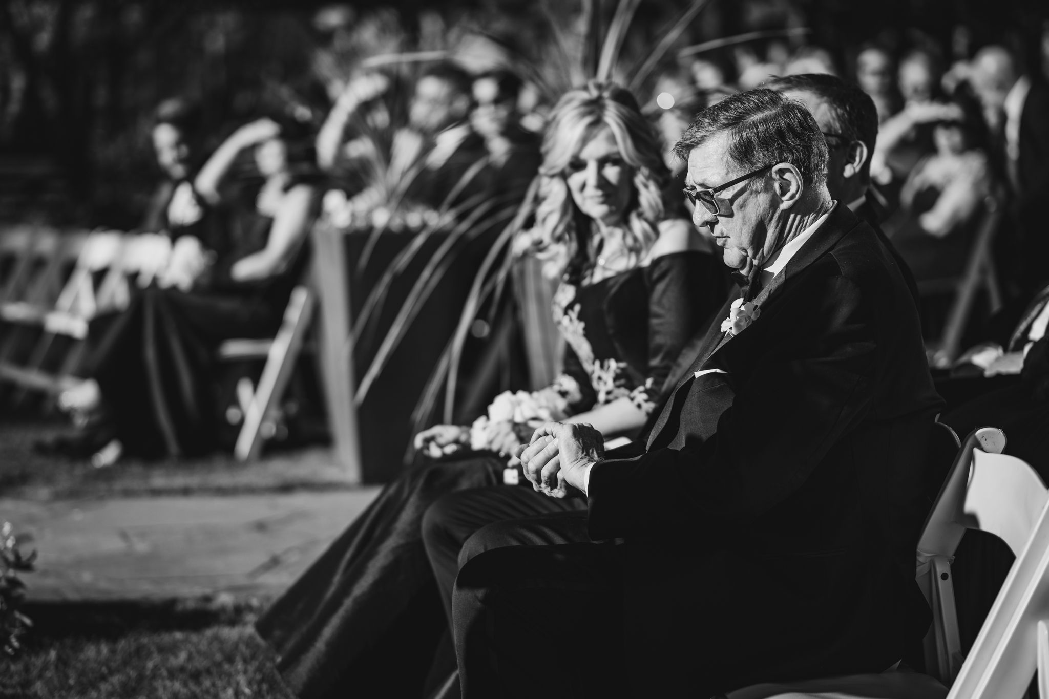 Wedding guests seated at an outdoor ceremony in black and white, with a man and woman in the foreground.