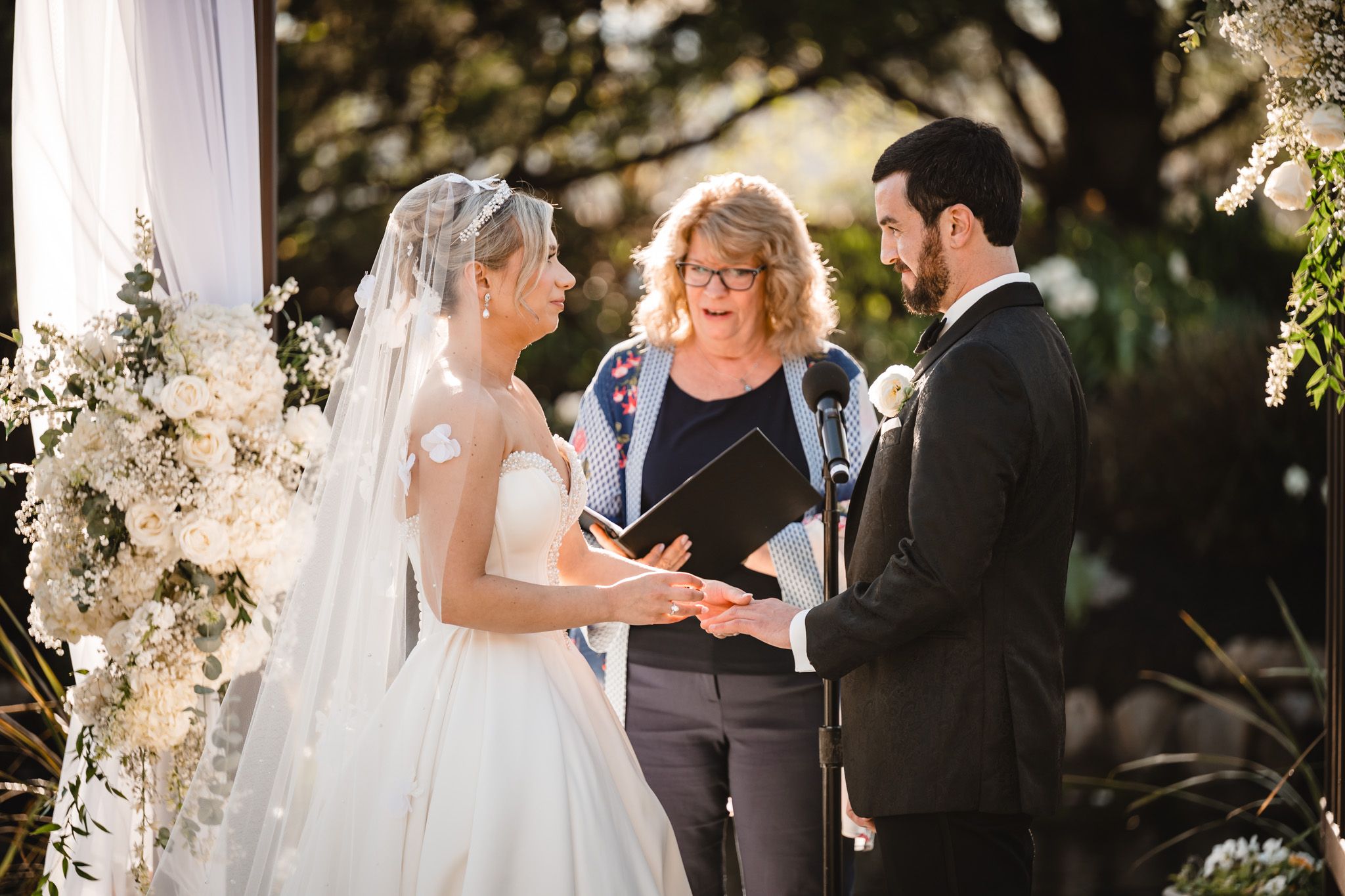 Bride places ring on grooms finger during outdoor ceremony with white floral arrangements nearby.