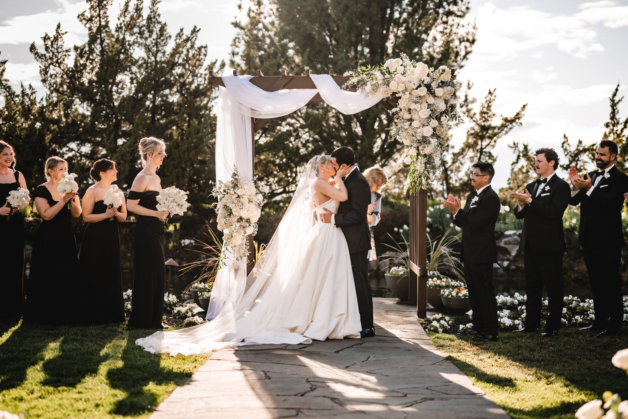 Bride and groom share first kiss under a draped floral arch flanked by wedding party in black attire.