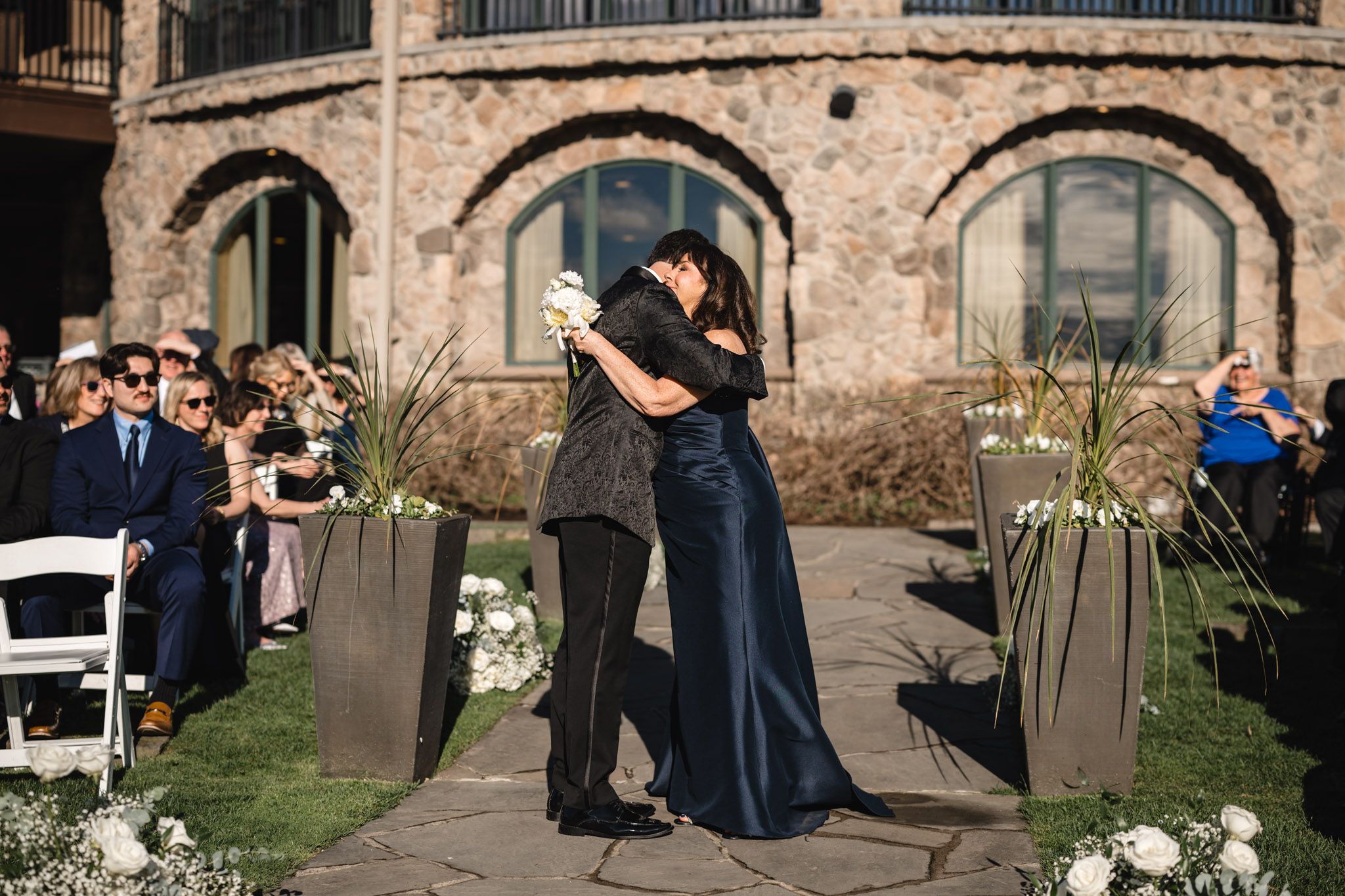 Two guests share an emotional hug along the outdoor ceremony aisle lined with white flowers.