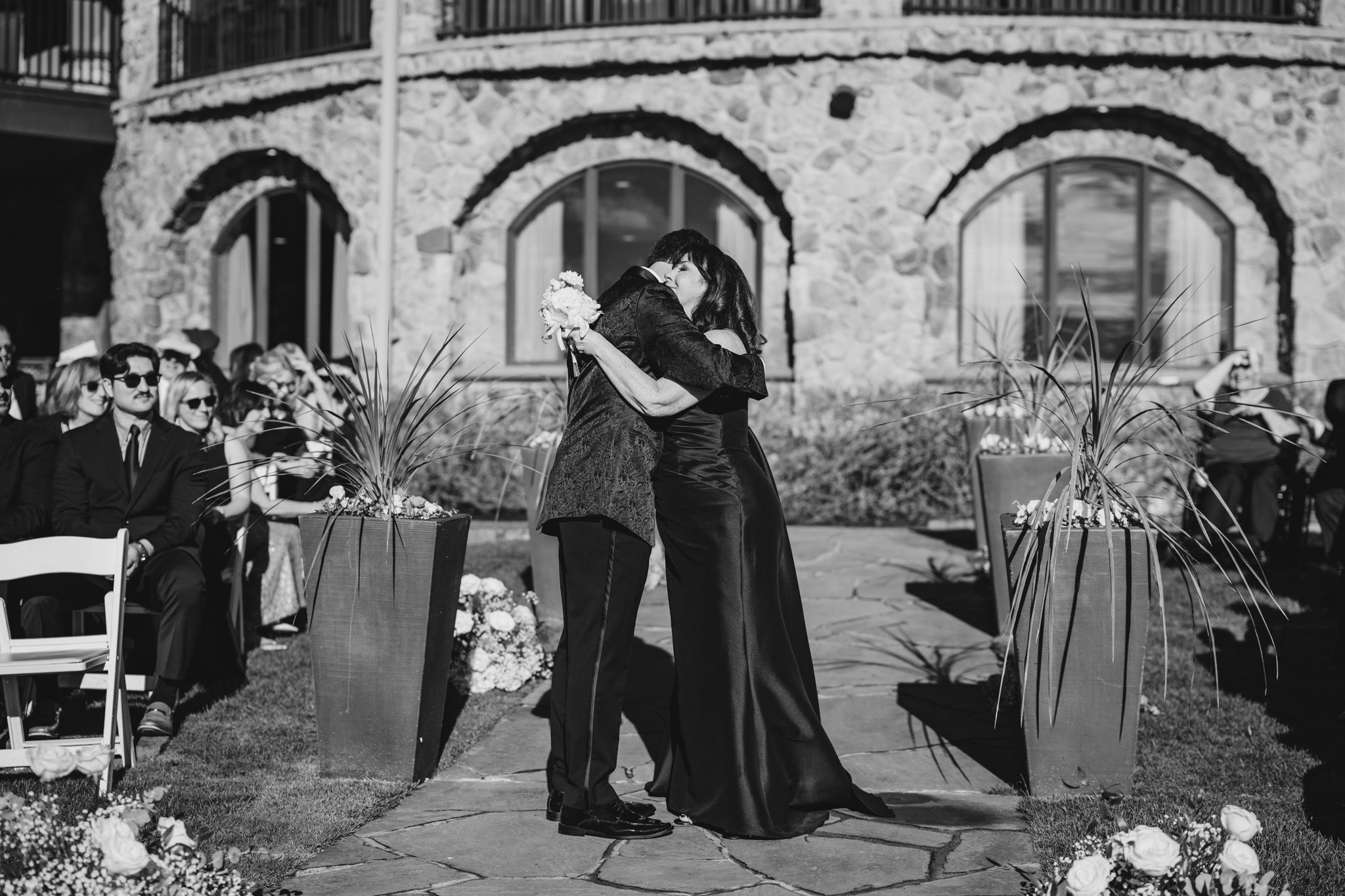 Black and white photo of two guests sharing an emotional embrace during an outdoor wedding ceremony.