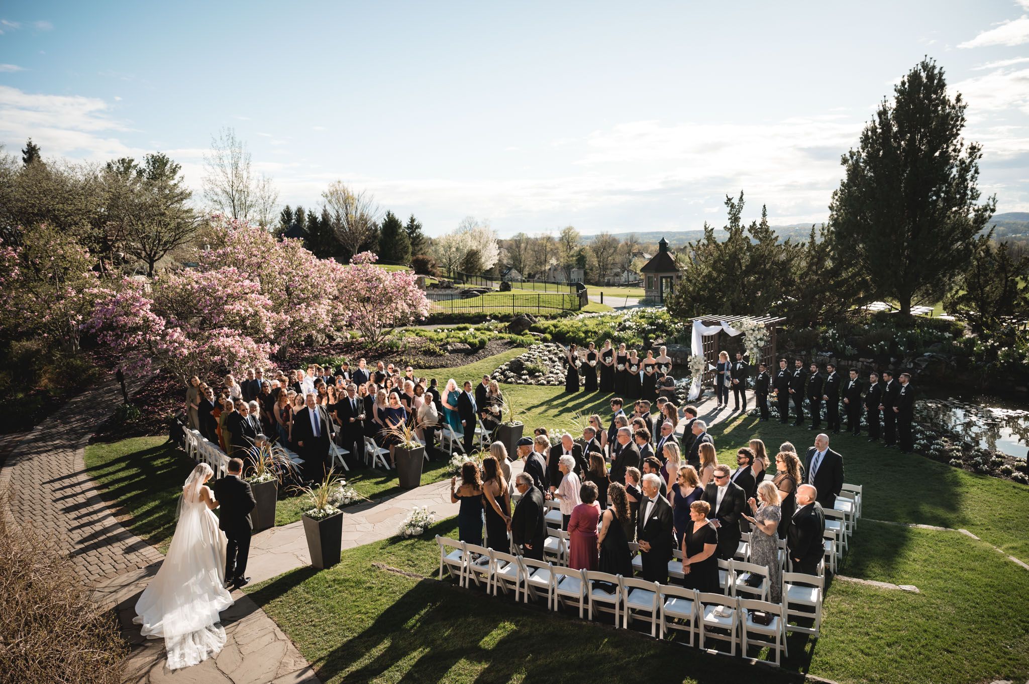 Outdoor spring wedding ceremony with guests seated on white chairs surrounded by blooming magnolia trees.