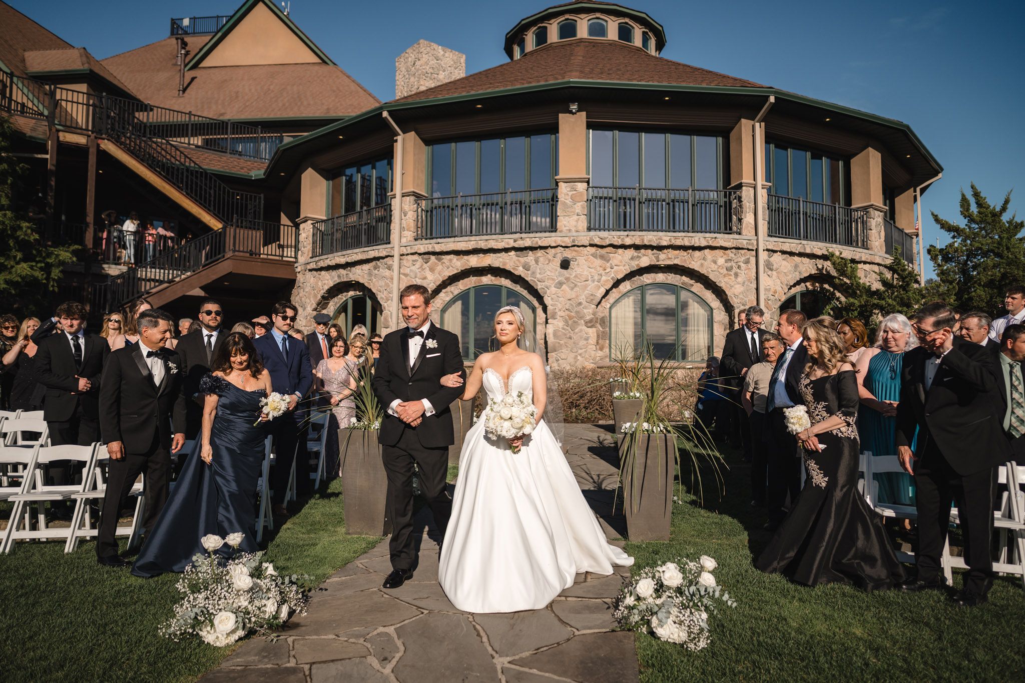 Bride and groom walk back up the aisle after outdoor ceremony in front of stone venue.