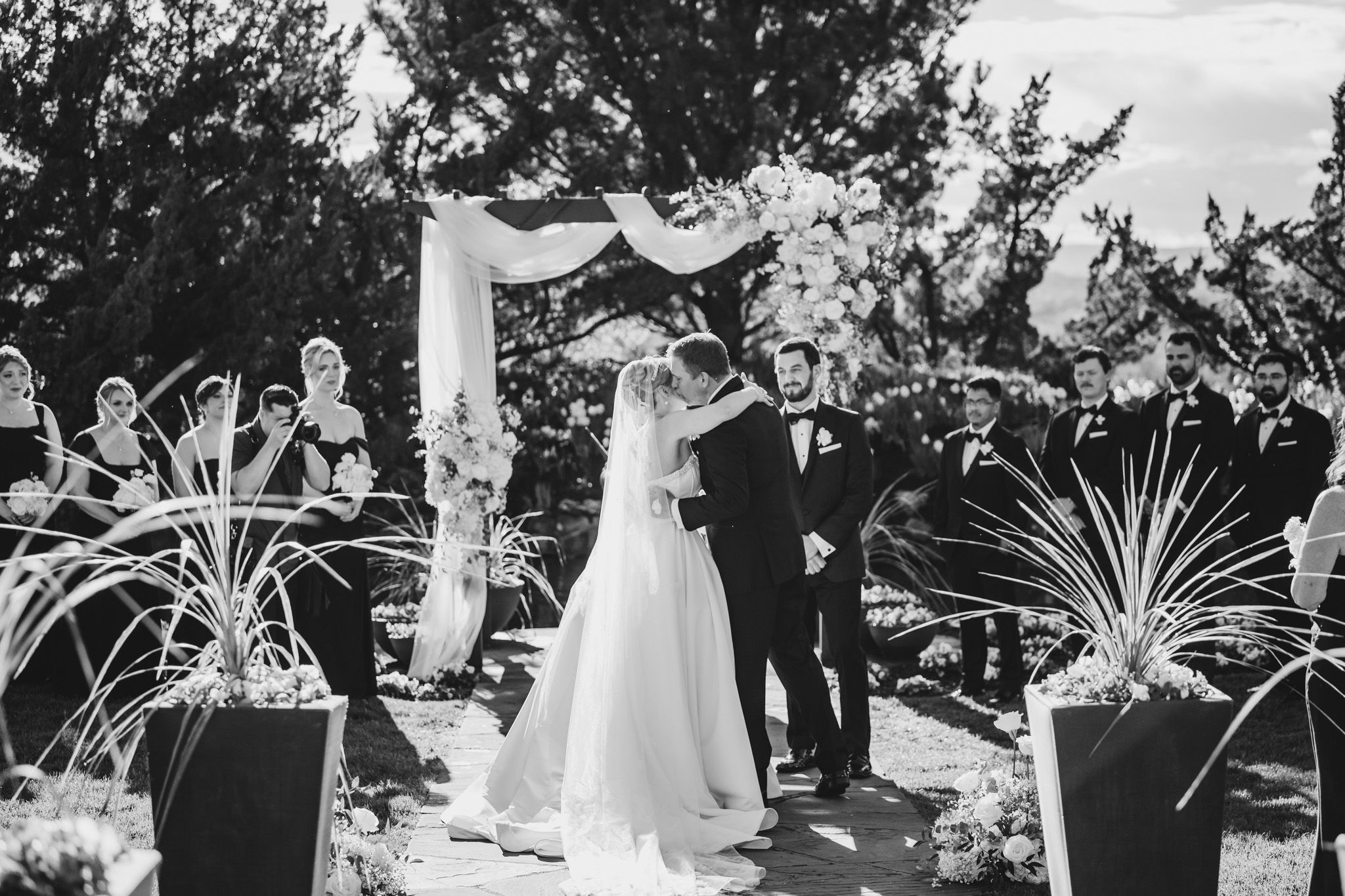Bride and groom share first kiss at outdoor wedding ceremony beneath floral arch with wedding party.