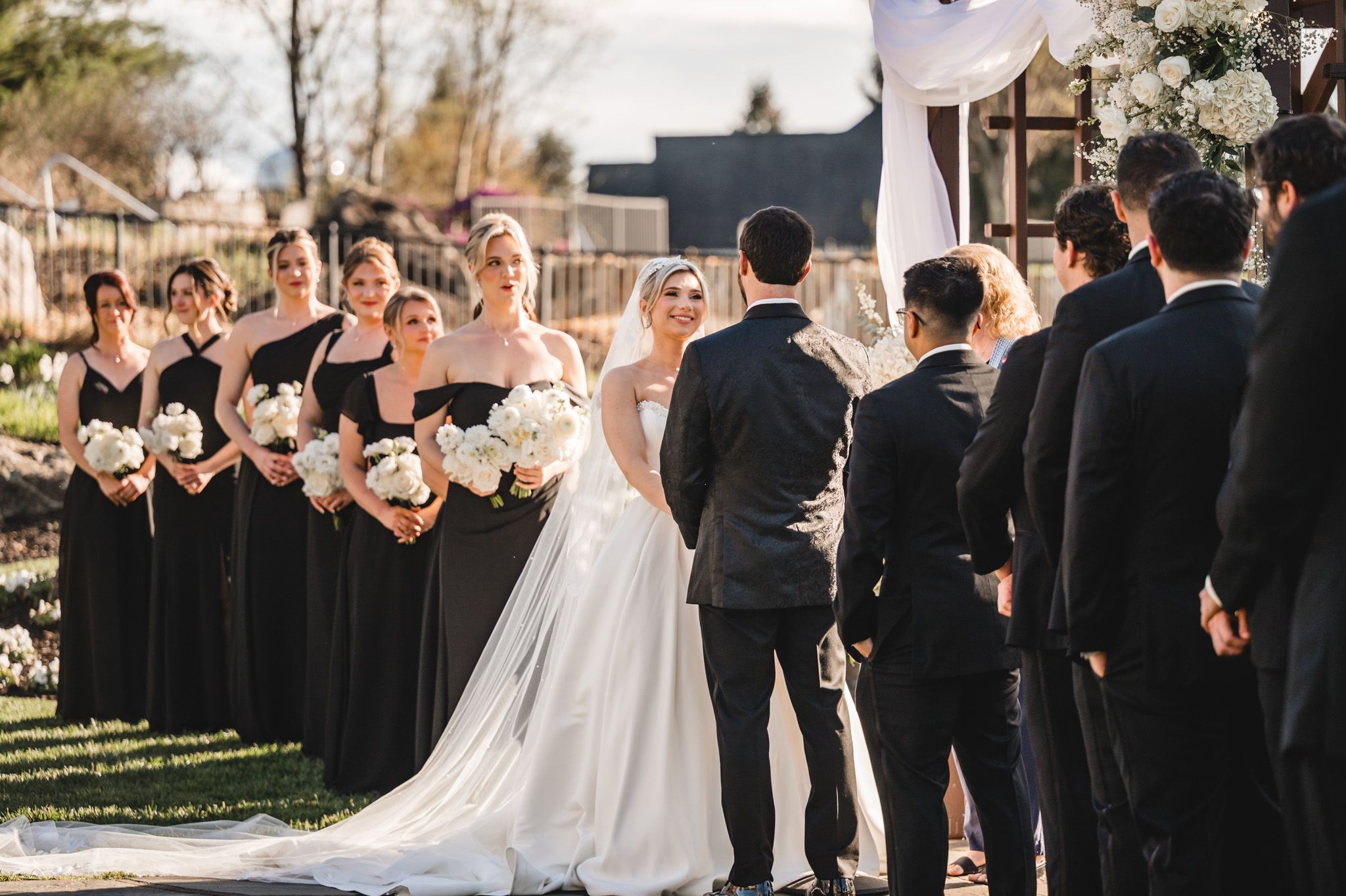 Outdoor wedding ceremony with bride in white gown, bridesmaids in black dresses holding white bouquets, and groomsmen in suits.