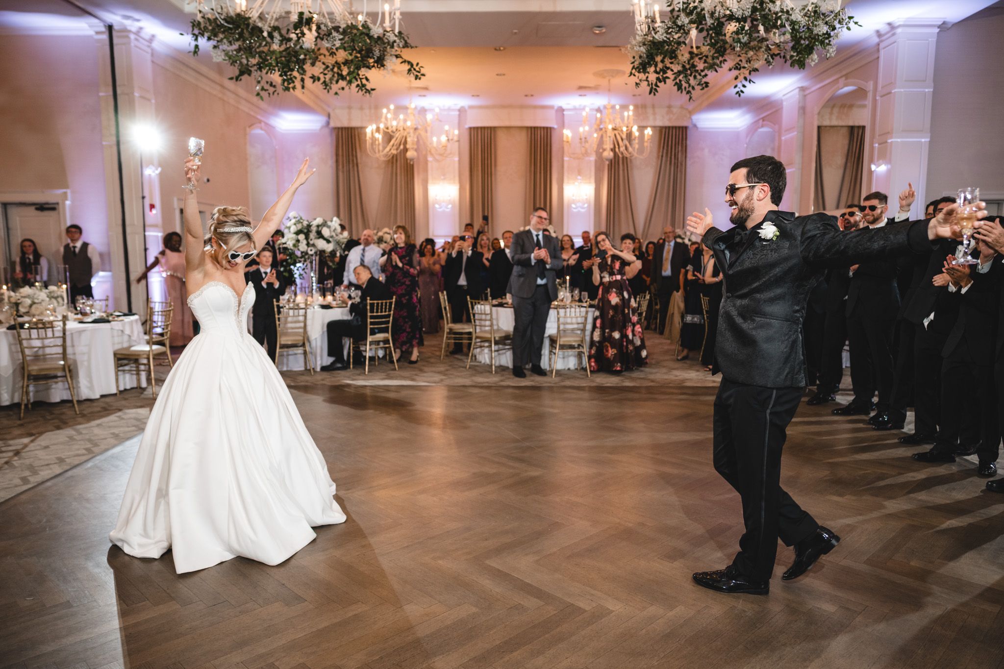 Bride and groom share their first dance on a herringbone wood floor surrounded by wedding guests.