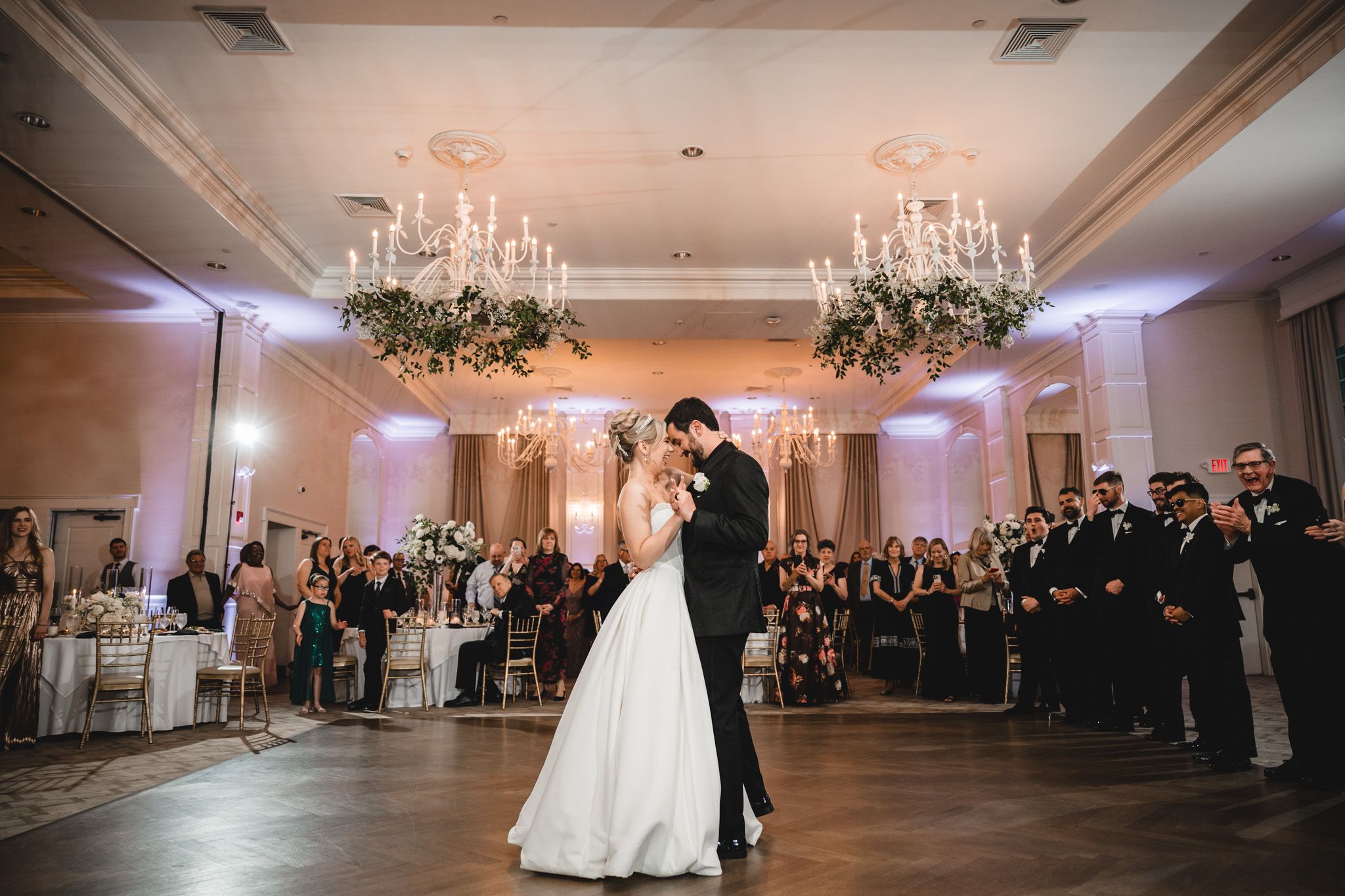 Bride and groom share first dance in elegant ballroom with chandelier and floral arrangements at wedding reception.
