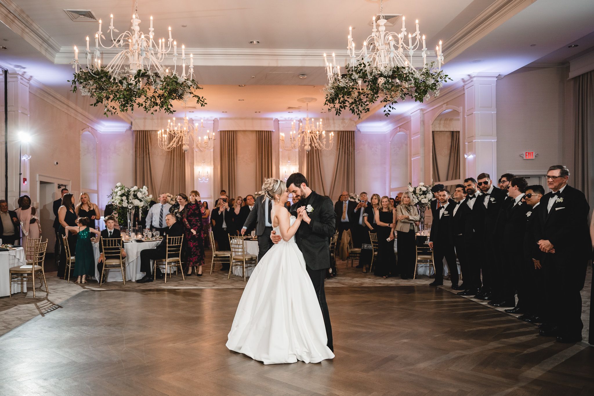 Bride and groom share first dance under chandelier as wedding guests look on in elegant ballroom.