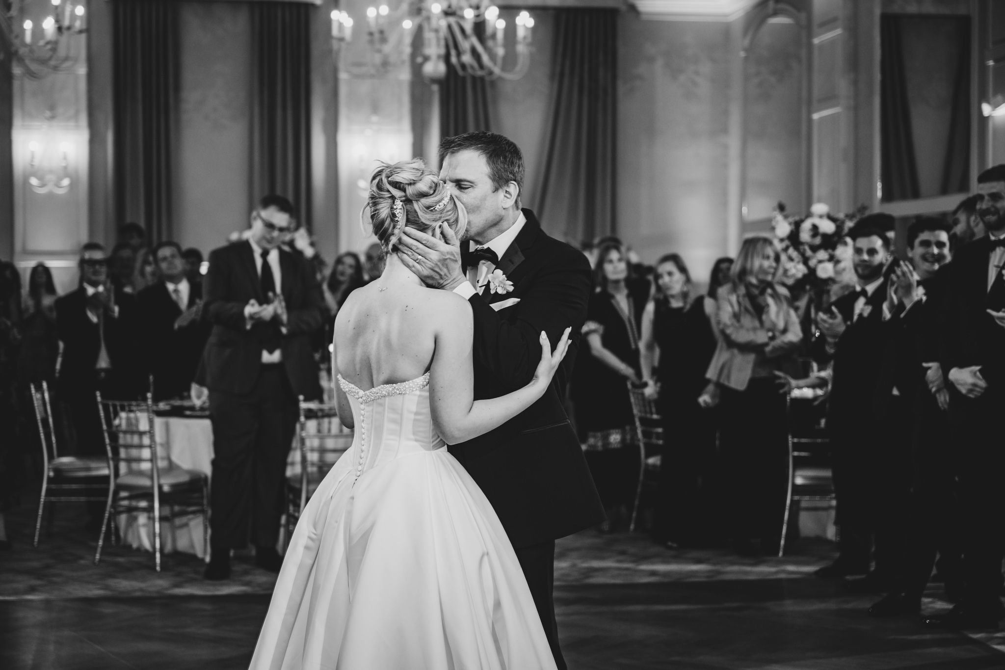 Bride and groom share their first dance in elegant black and white wedding photo with guests applauding.