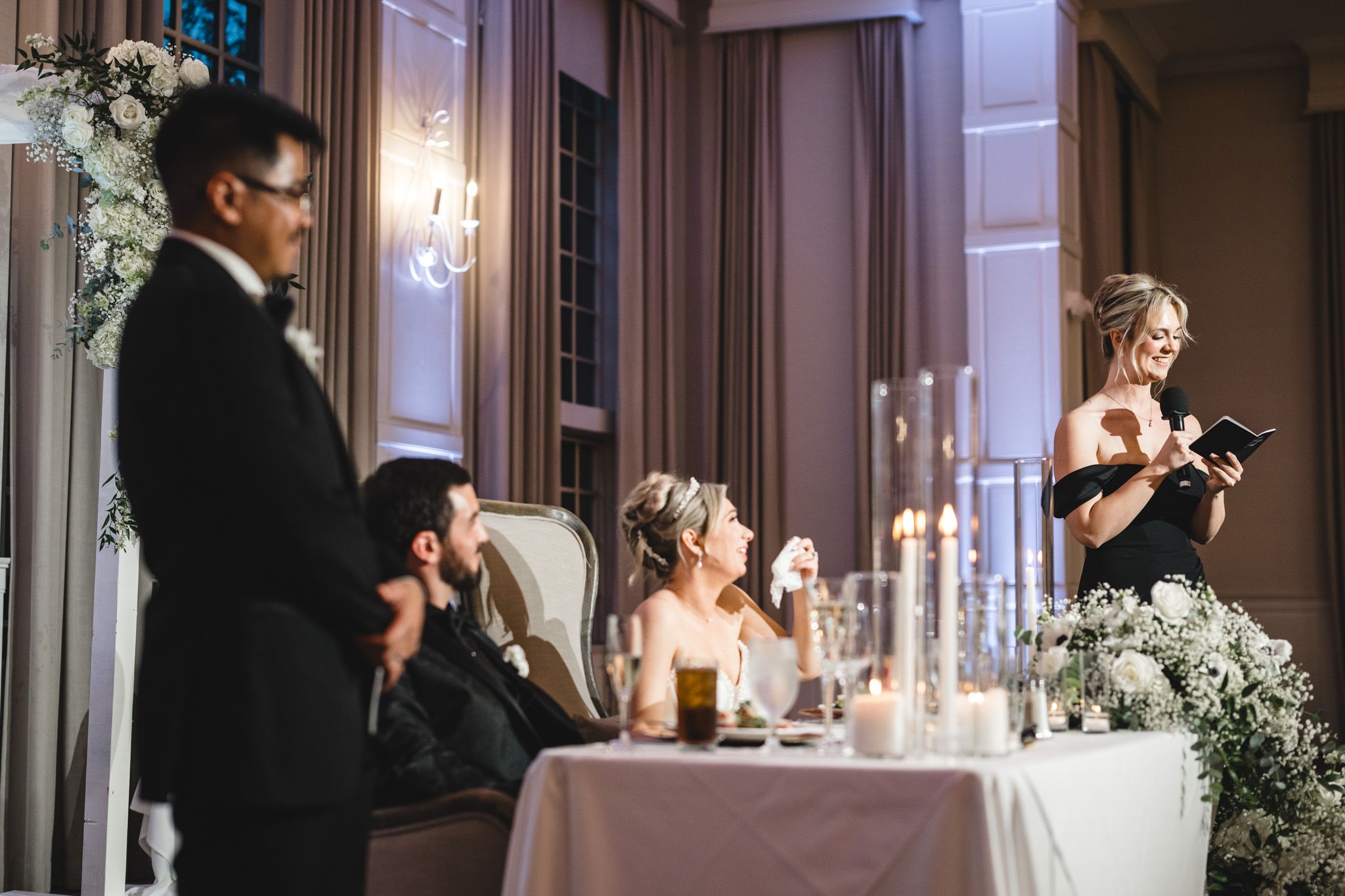 Wedding guest delivers heartfelt speech at elegant candlelit reception while bride smiles at sweetheart table.