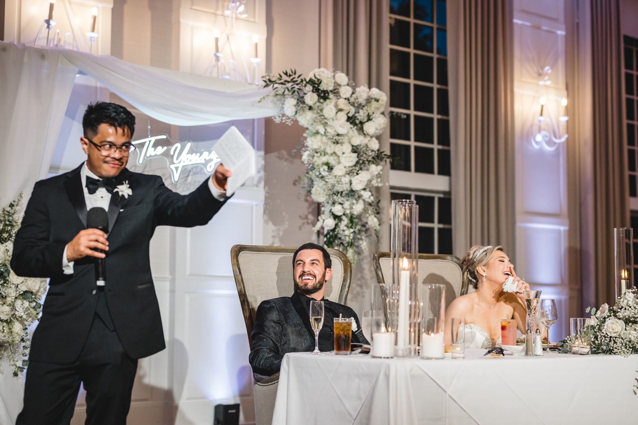 Best man giving a toast speech at wedding reception while bride and groom smile at sweetheart table with floral decor.