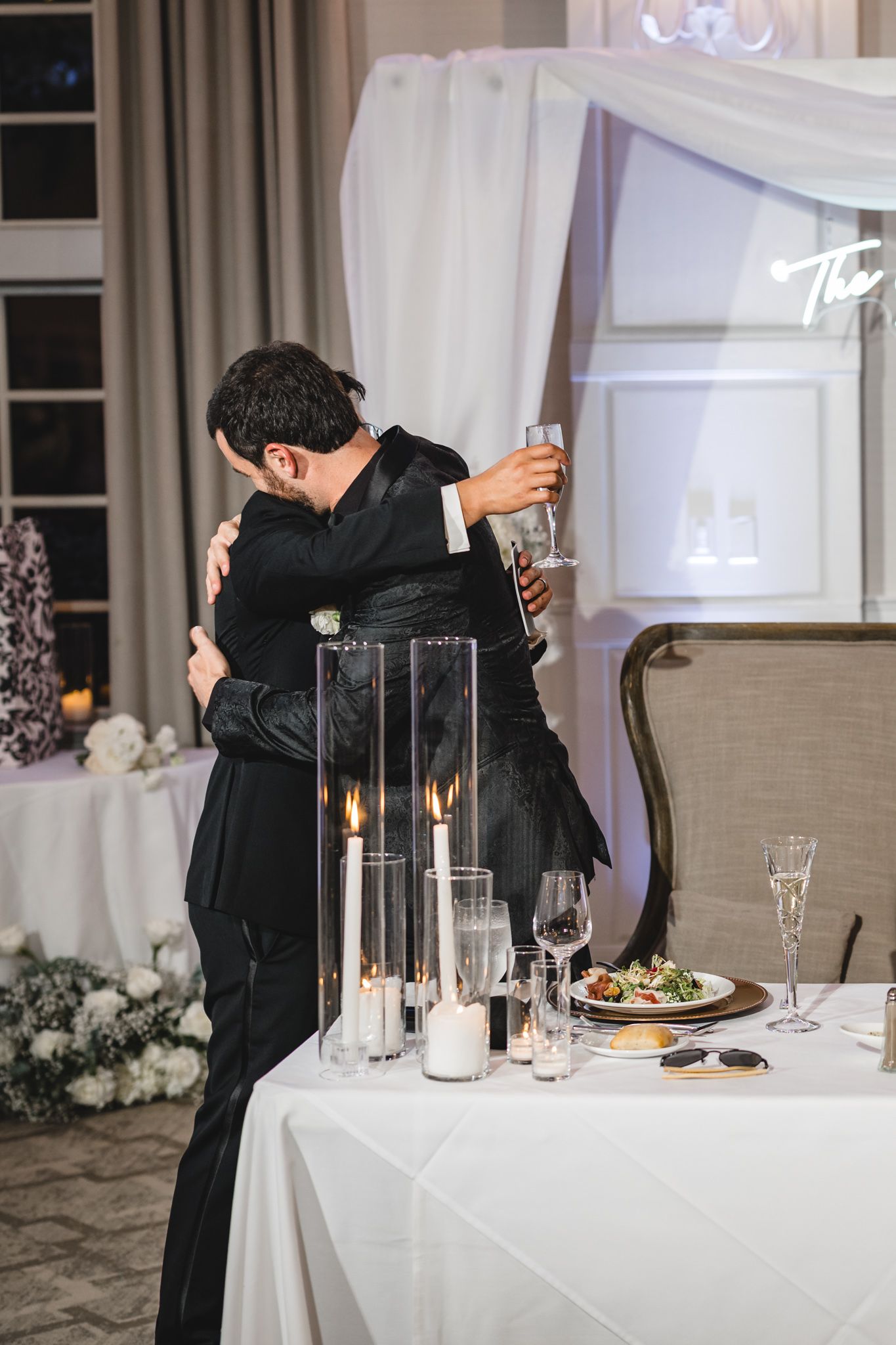 Two guests in black formal attire share a warm hug while holding drinks at an elegant candlelit wedding reception.