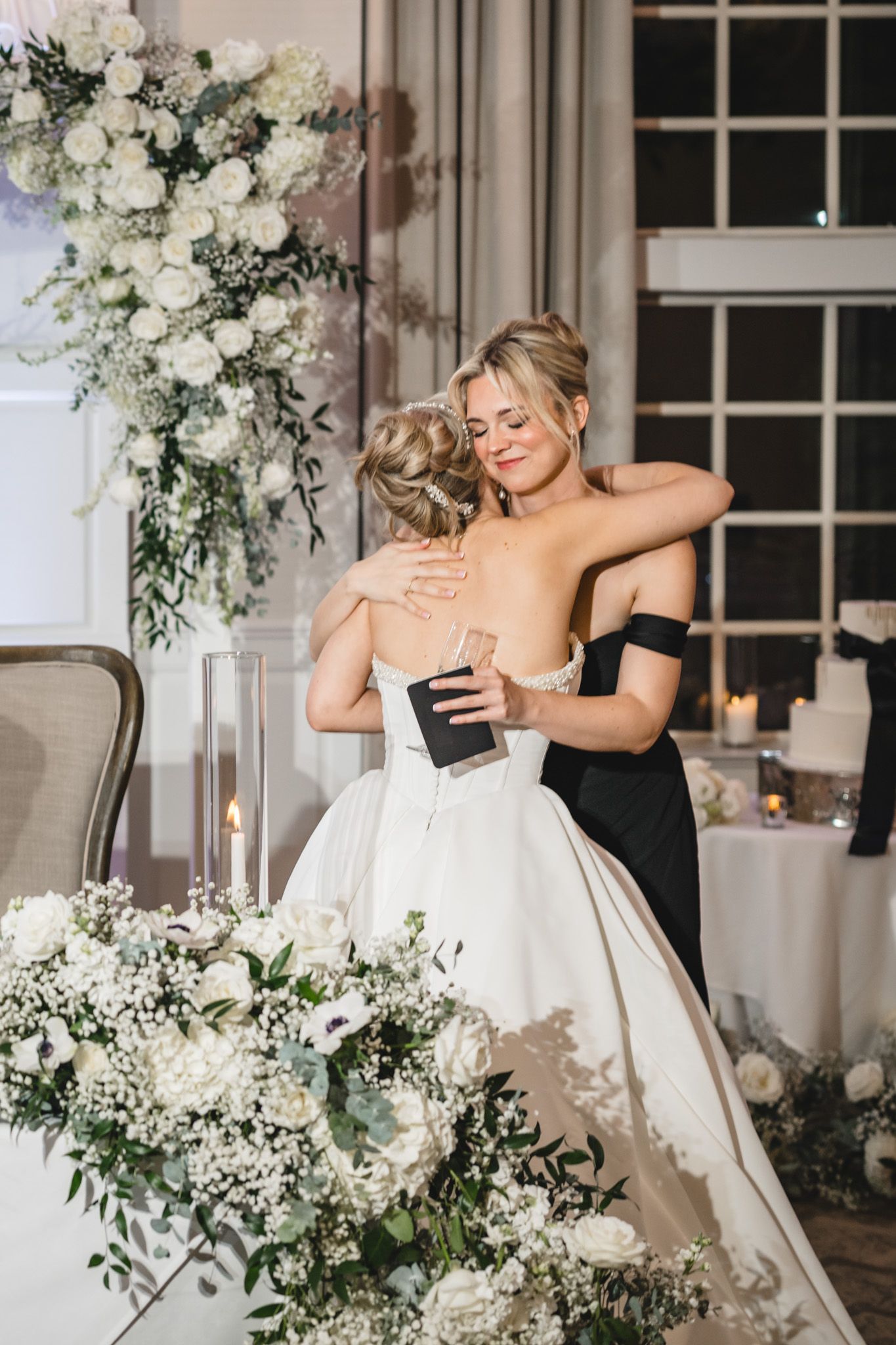 Bride and bridesmaid share emotional hug at wedding reception surrounded by lush white floral arrangements.